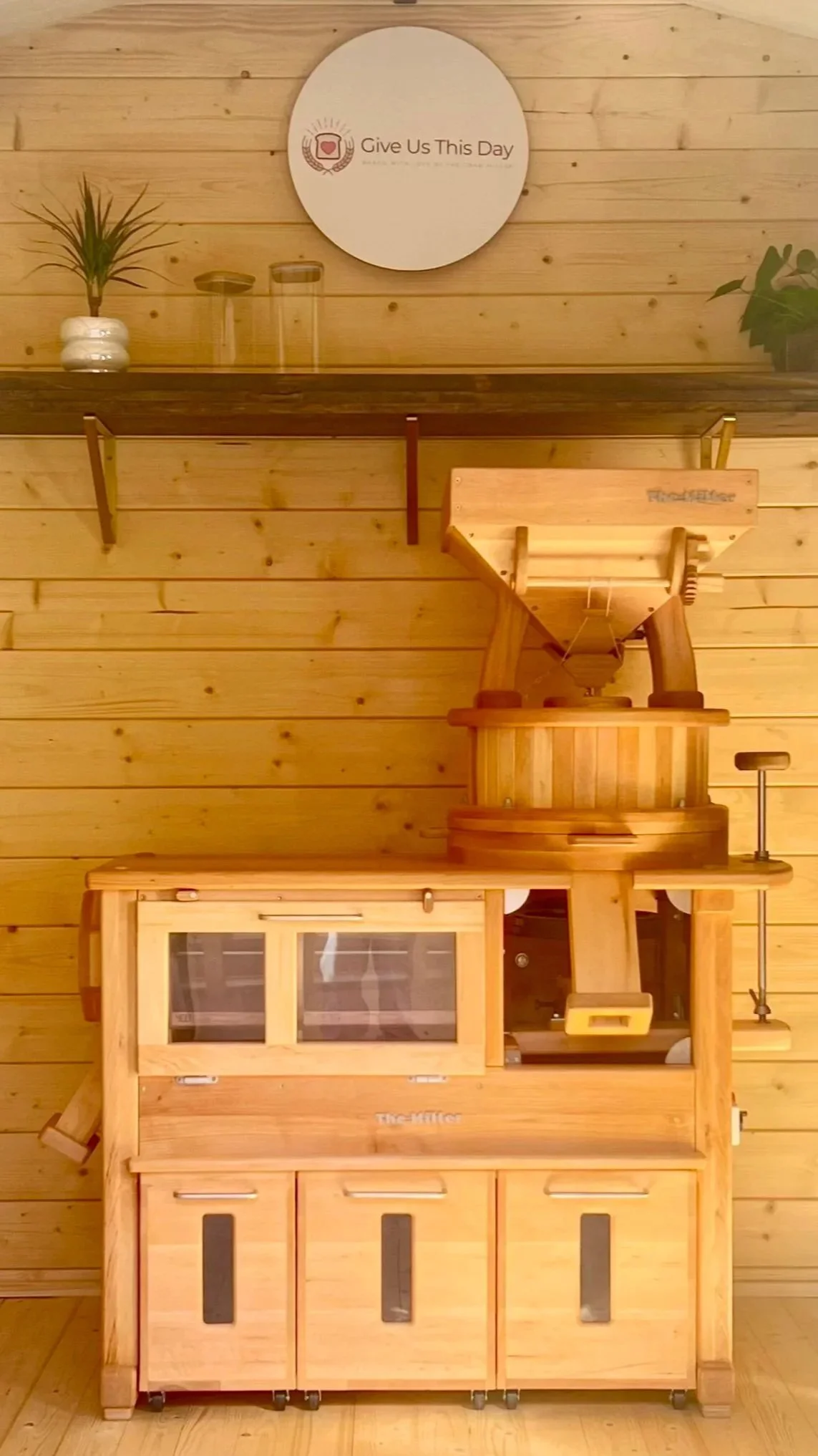 A wooden dough sheeter machine used for rolling dough, placed in a room with wooden panel walls and a wooden shelf above it. The room has plants and a sign that says 'Give Us This Day' on the wall.