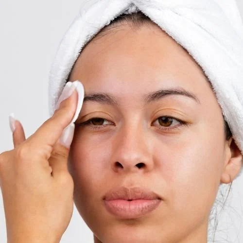 Woman with a towel wrapped around her head gently dabbing her eyebrow area with a cotton pad, demonstrating how to cleanse brows carefully during the first 48 hours after a nano brow procedure.