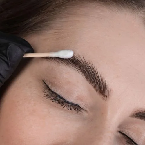 Close-up of a person applying a thin layer of ointment to their brows with a clean cotton swab, illustrating how to lightly moisturize nano brows during the first week of healing.
