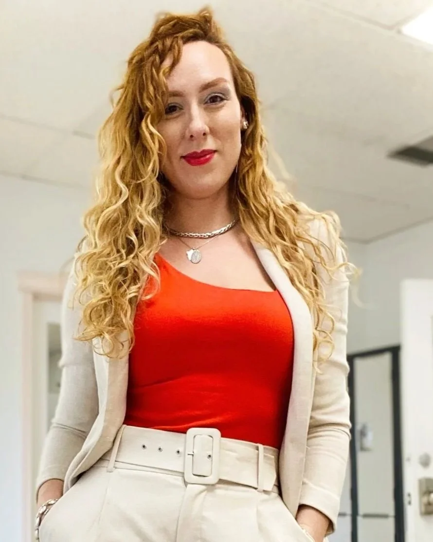 Alexandra with long curly red hair wearing a red top, beige blazer, and beige belt, smiling in an indoor setting.