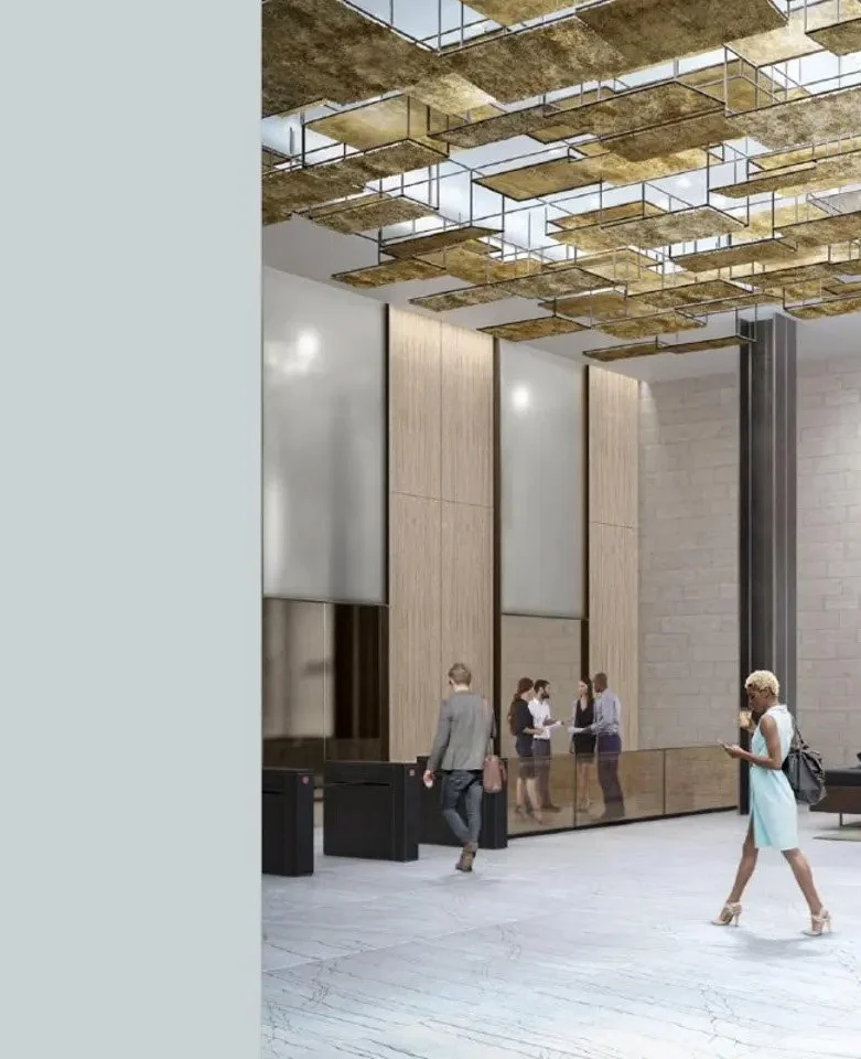 Interior of a modern lobby with a high ceiling decorated with gold-colored rectangular panels, and a reception desk where several people are working. Other individuals are walking or standing, one woman in a light blue dress walking across the space.