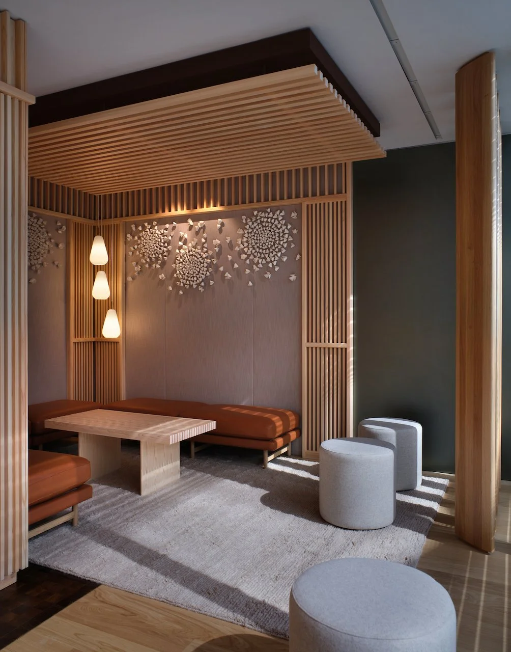 A modern interior lounge area featuring wooden slats on the ceiling and walls, with a wall decoration of white butterfly sculptures. There are three wall-mounted pendant lights, a low wooden table, upholstered seating in brown and white, and a striped rug on the floor.