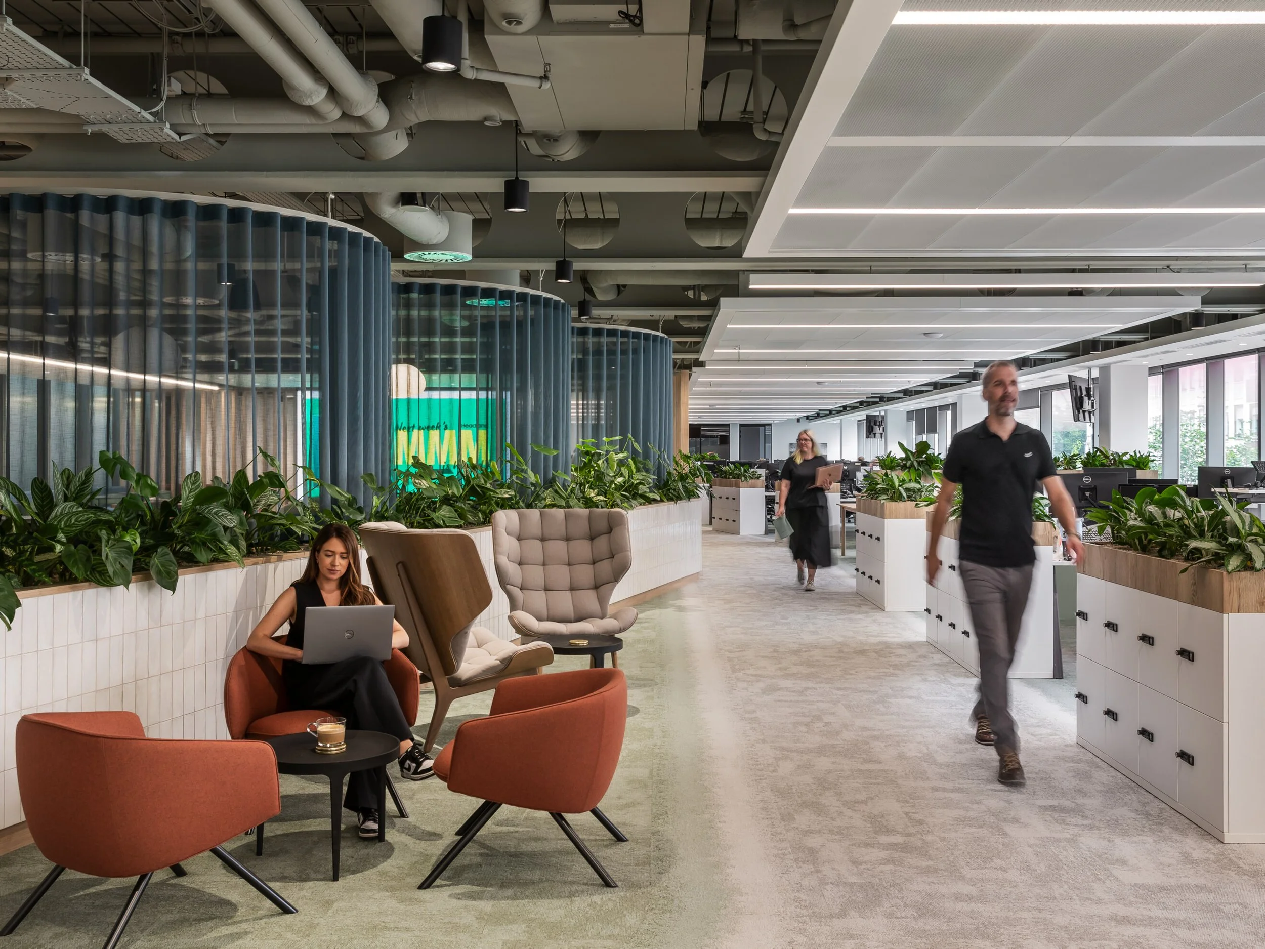 Modern office space with lounge area, plants, and employees walking and working.