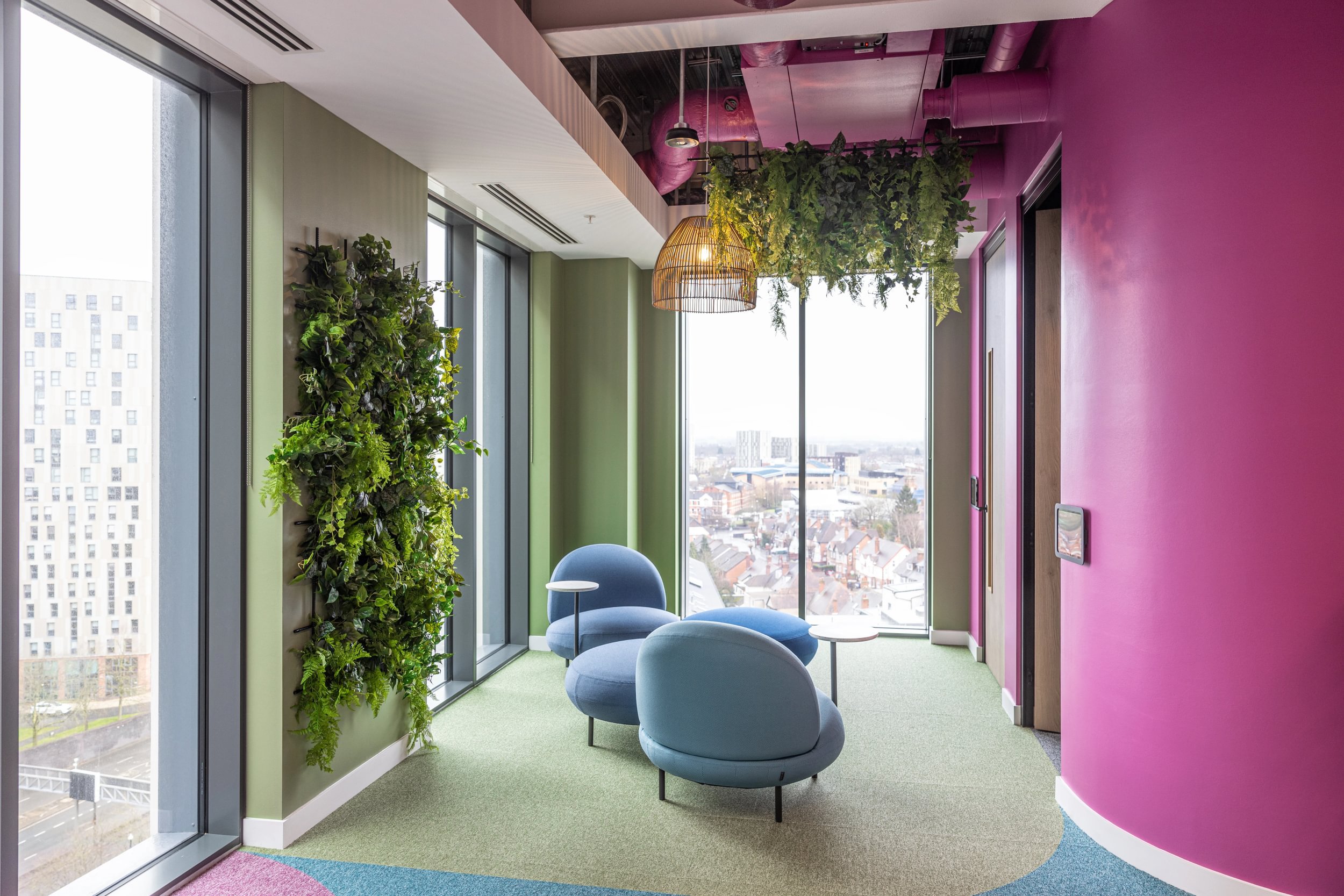 Modern office corner with colorful walls, green and pink, featuring a vertical garden, two blue lounge chairs, small white tables, large window with city view, and hanging light fixtures with greenery.