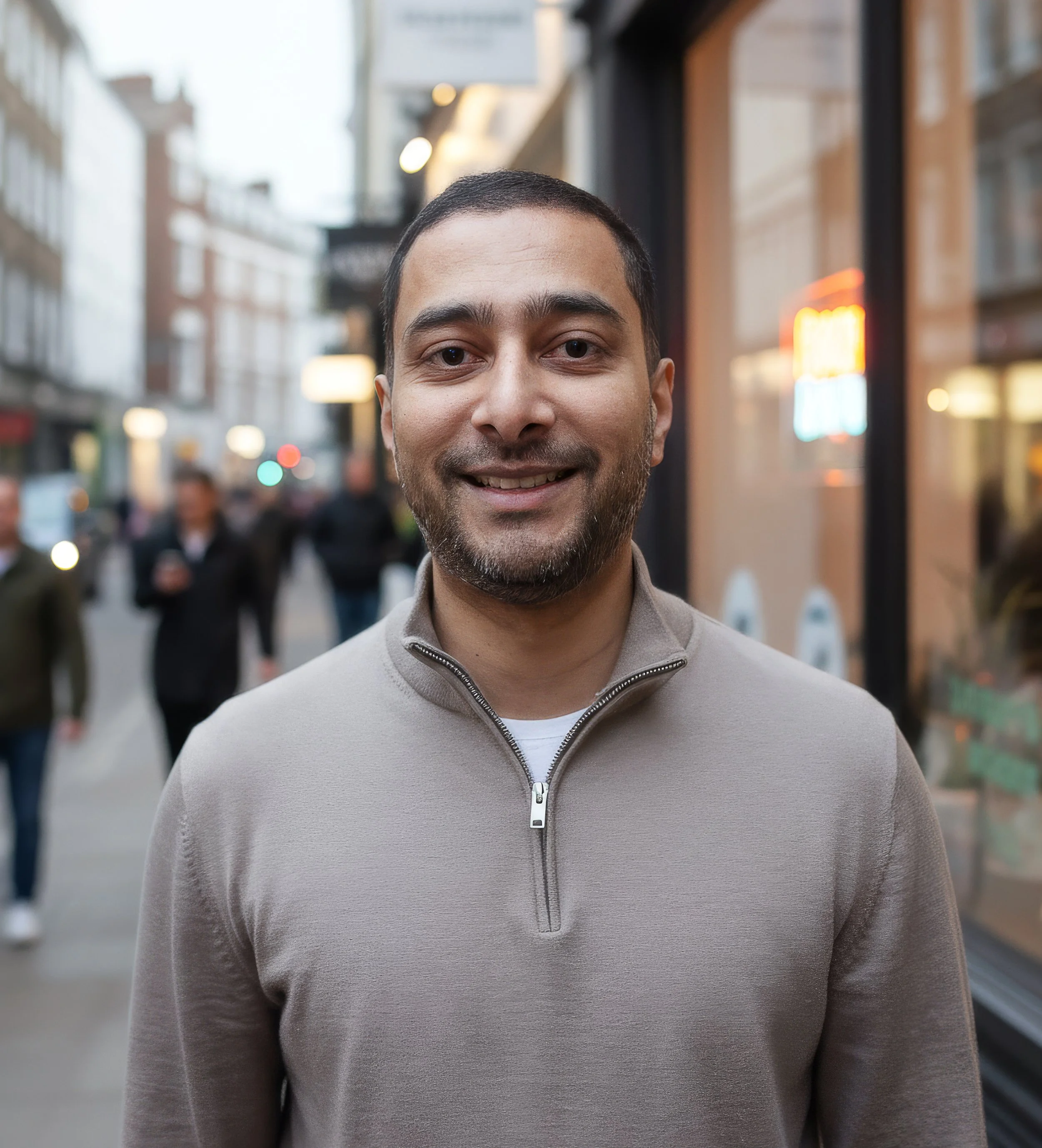 Smiling man standing on a busy city street with storefronts and pedestrians in the background.