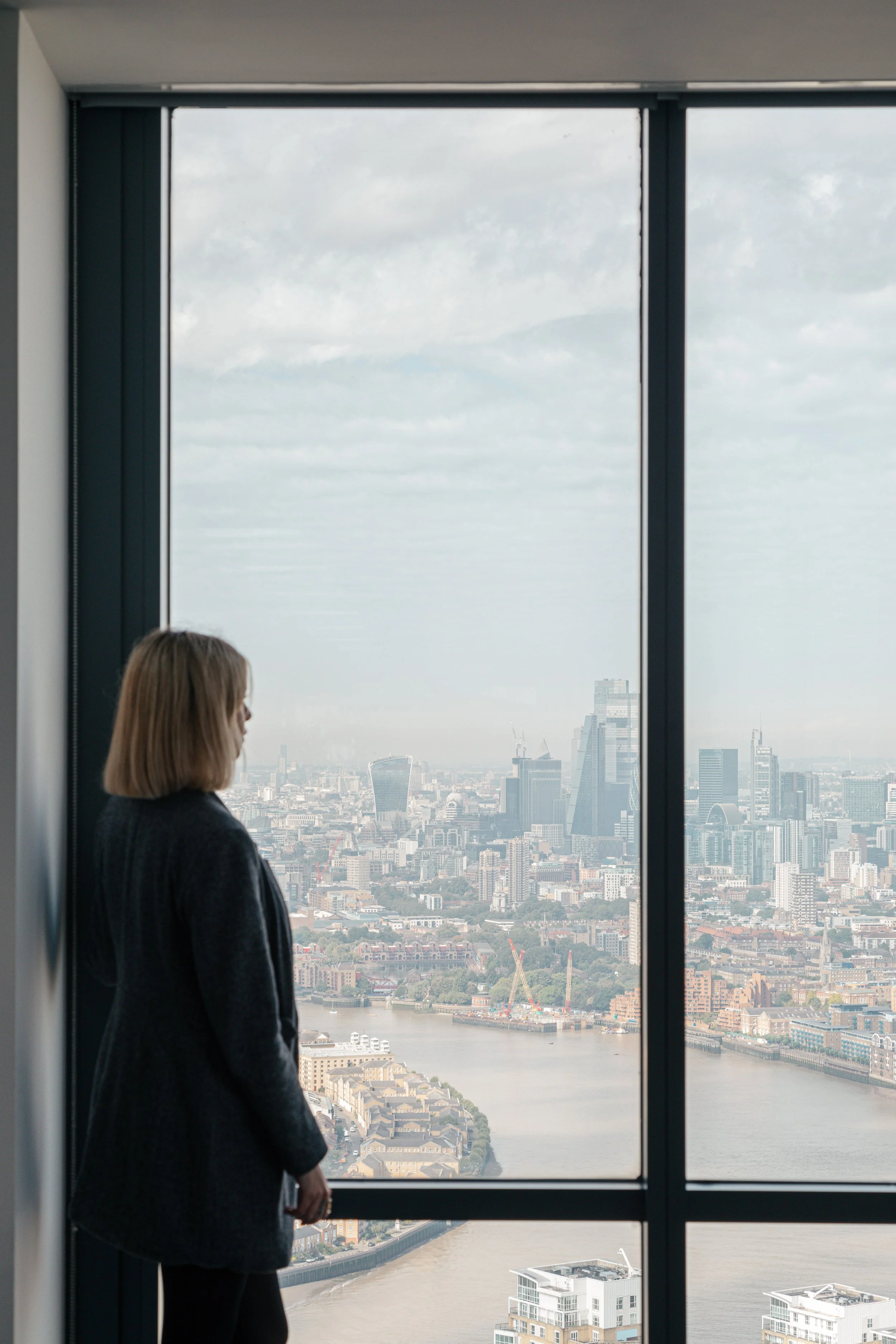 A woman with shoulder-length hair standing indoors near a large window, overlooking a city skyline with tall buildings and a river below.