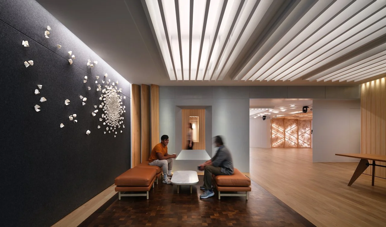 Two men sit on orange cushioned benches facing each other in a modern, well-lit lounge area, with a woman walking past in the background. The room features wood, gray, and white design elements with a decorative wall art installation on the left and a light-colored wooden floor.