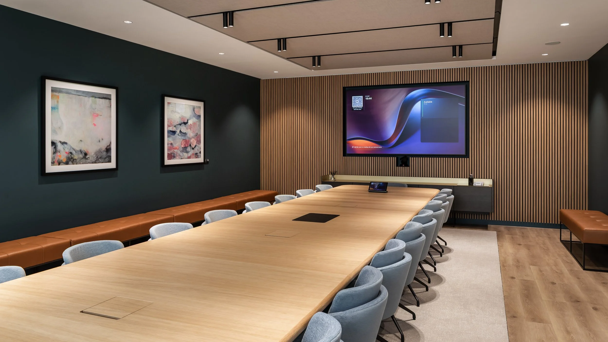 A modern conference room with a large wooden table surrounded by light gray chairs, a large wall-mounted screen displaying a digital interface, and art paintings on a dark green wall, with wood-paneled accents and leather benches along the wall.