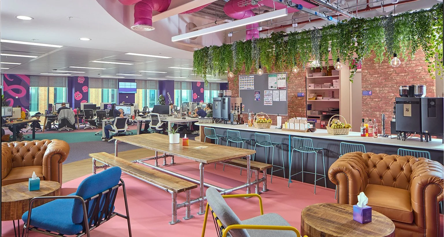 Bright modern office lounge area with colorful decor, leather and fabric chairs, a wooden table, a bar counter with barstools, and an open workspace in the background. Decor includes hanging plants, exposed brick wall, and pink ceiling pipes.
