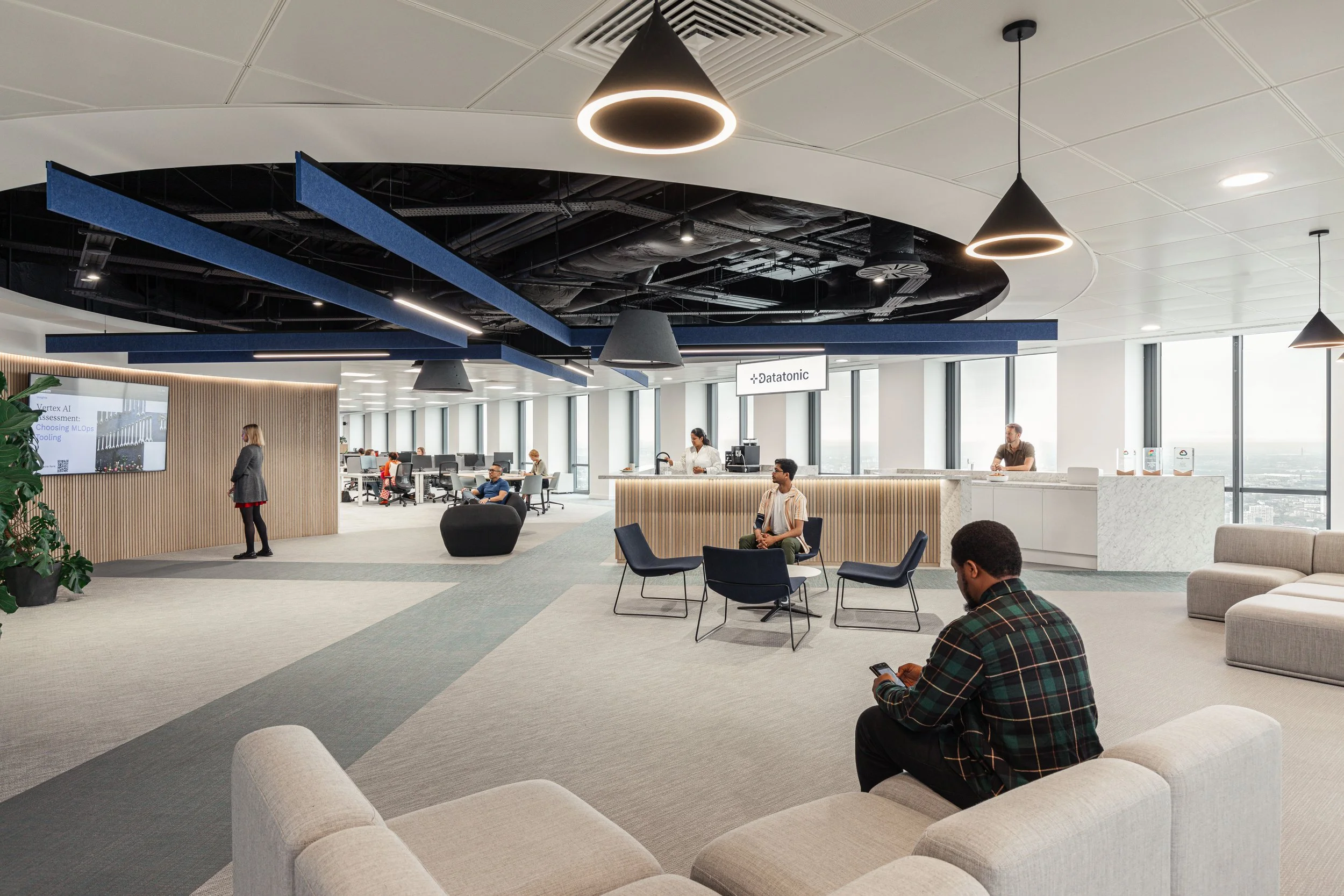 Modern office reception area with seating, large windows, and a reception desk. People are sitting and walking, with a woman at a presentation screen on a wooden wall. There are decorative ceiling elements and pendant lights.