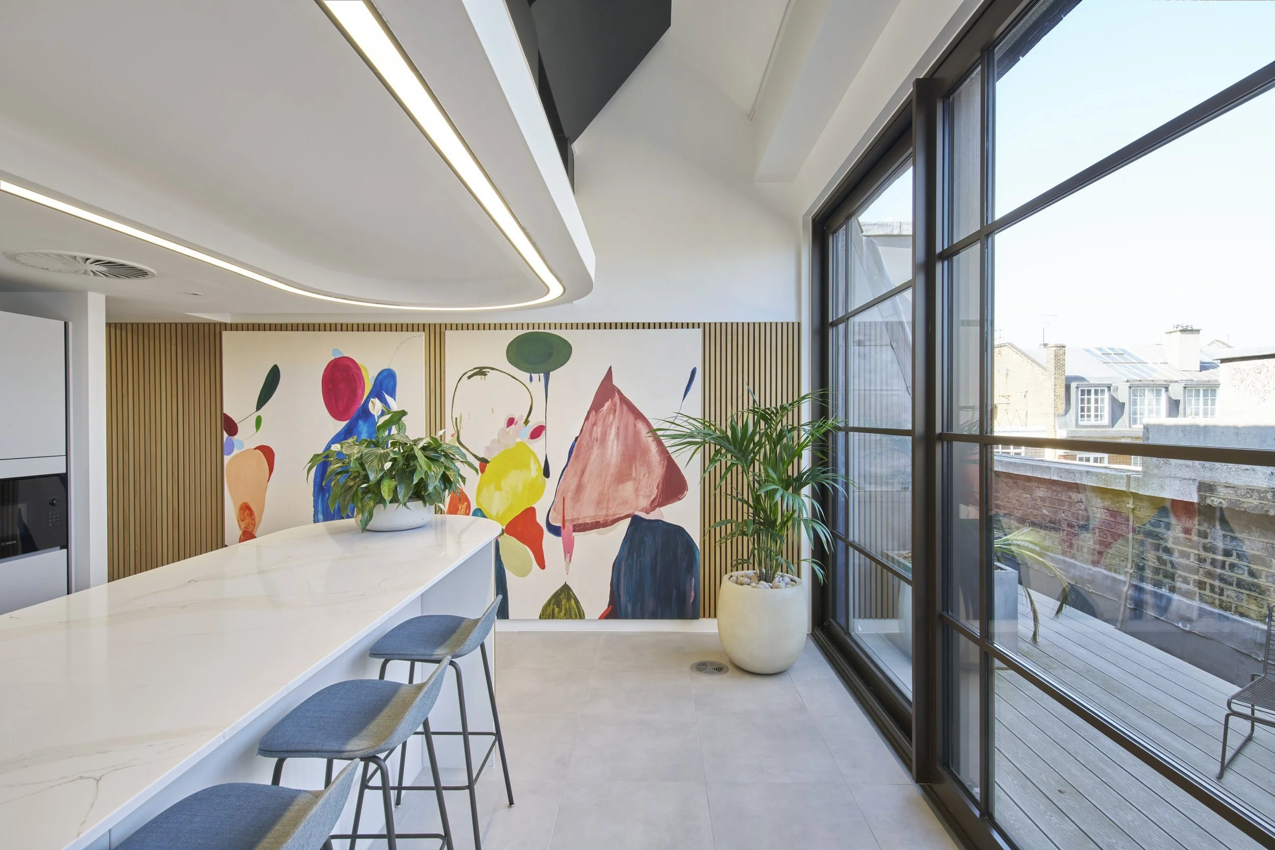 Modern kitchen with white marble island, blue cushioned bar stools, large abstract artwork, potted plants, and floor-to-ceiling glass windows overlooking balcony and neighboring rooftops.