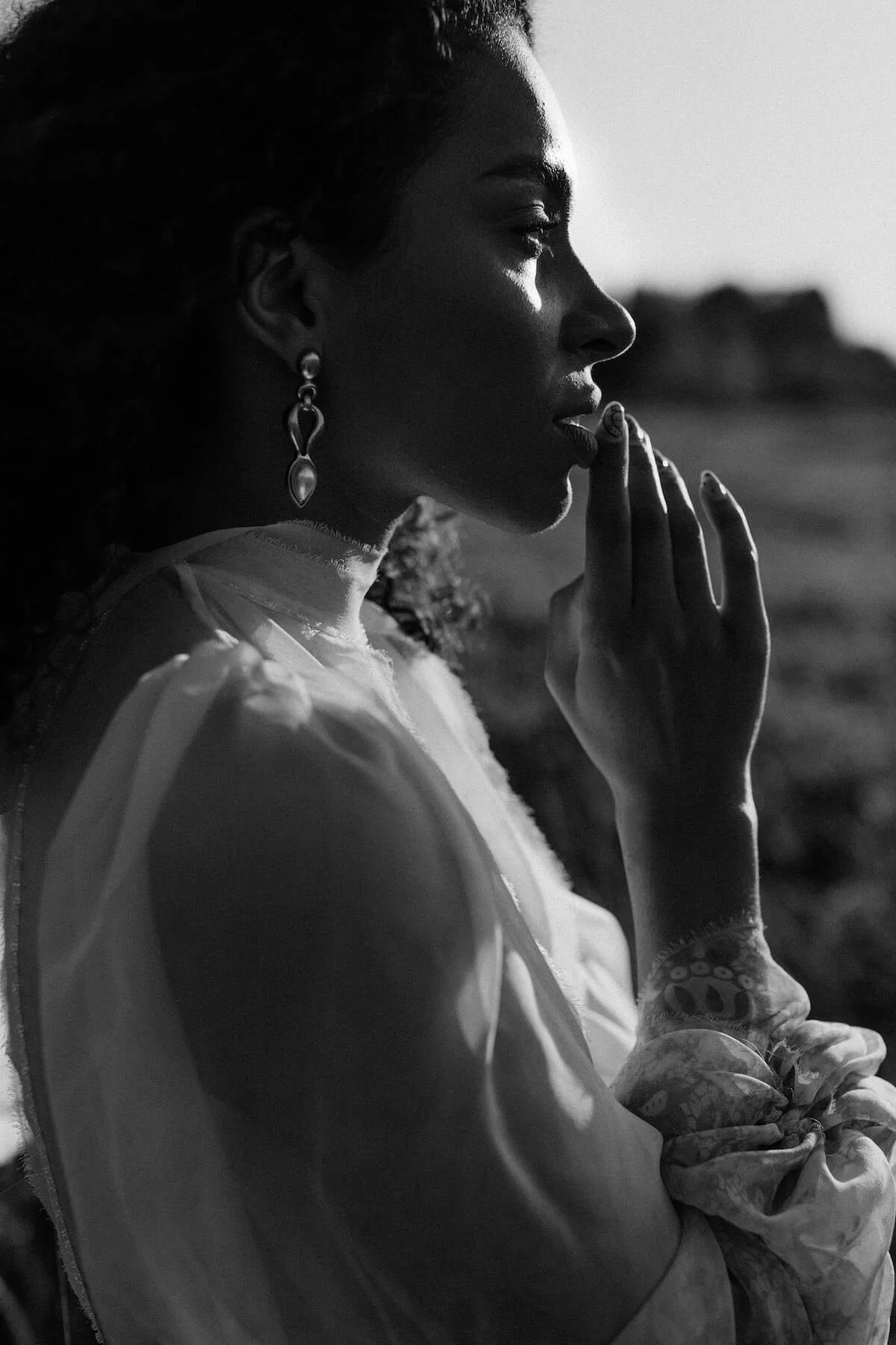 A black and white profile photo of a woman gazing into the distance, with her hand near her lips, wearing earrings and a long-sleeve blouse, outdoors during sunset.