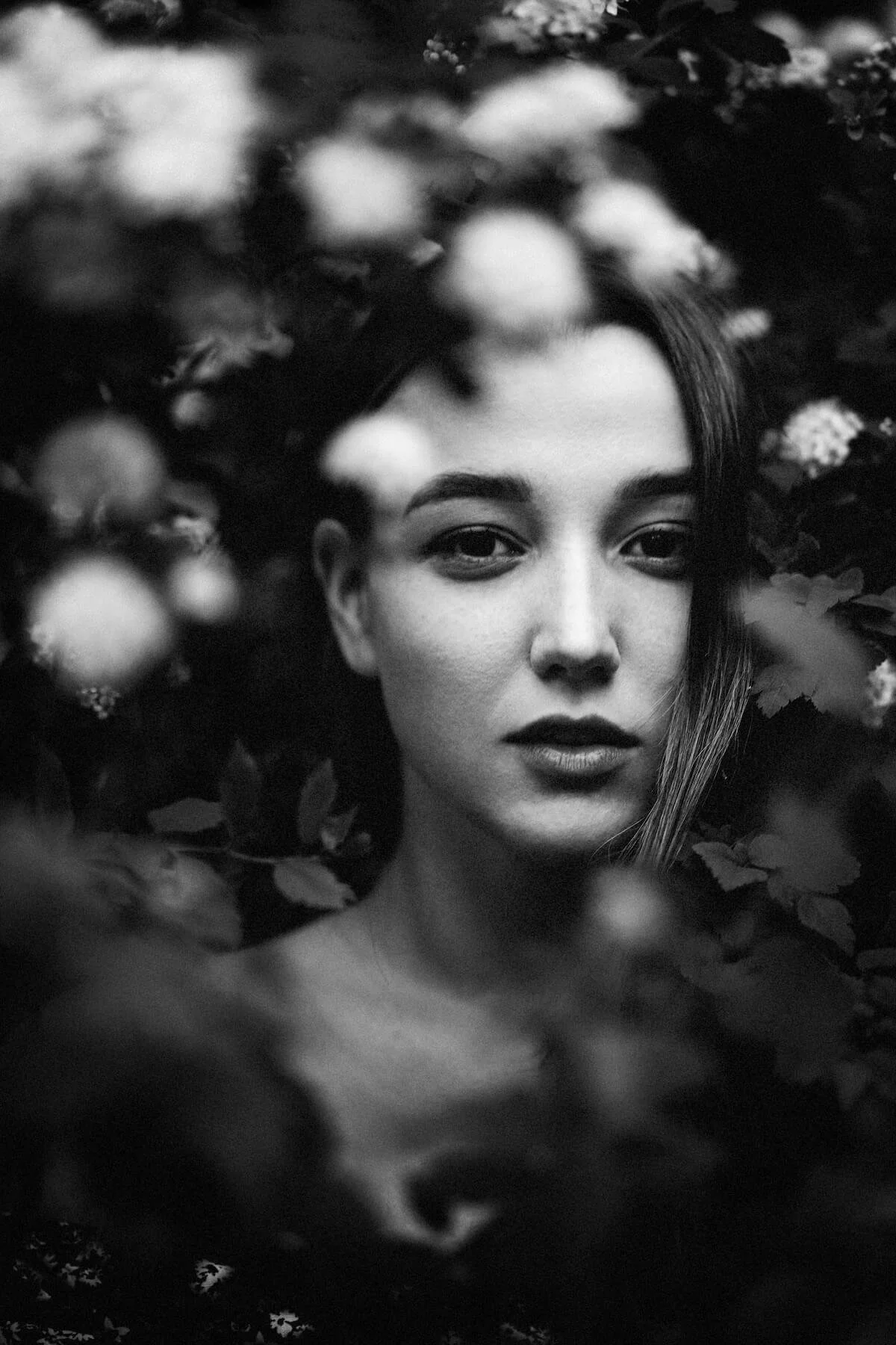 A black and white portrait of a woman with long hair, surrounded by blurred foliage and flowers.