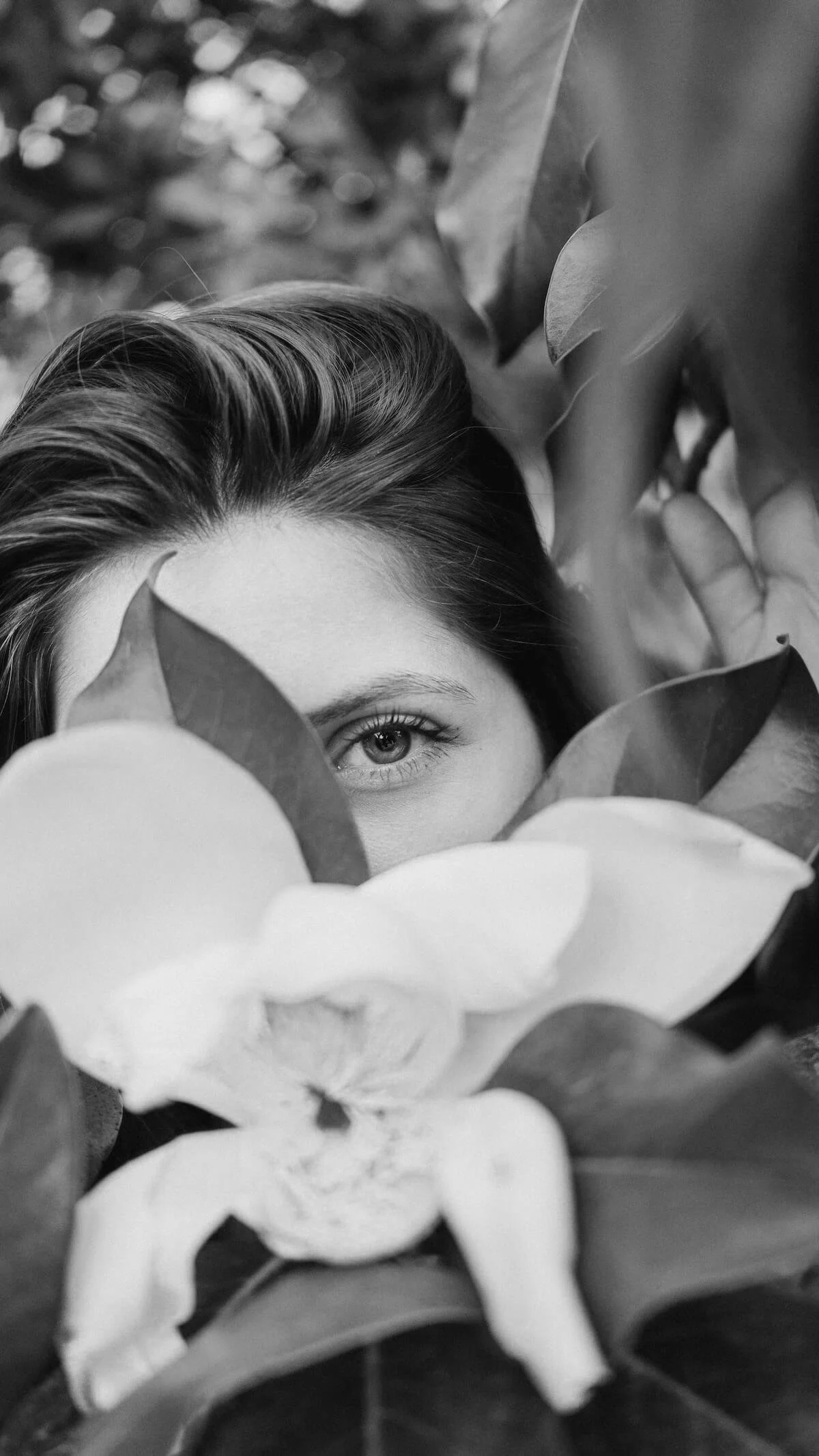 A woman partially hidden behind large leaves and flowers, with only one eye visible, in black and white.