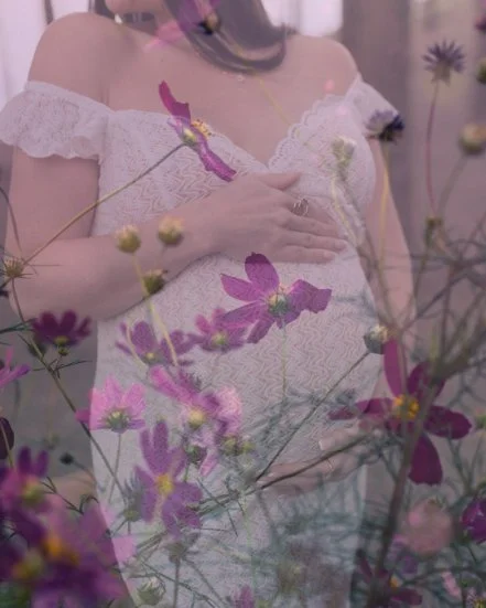 A pregnant woman in a white lace dress with off-the-shoulder sleeves, holding her belly with one hand while surrounded by pink and purple flowers.