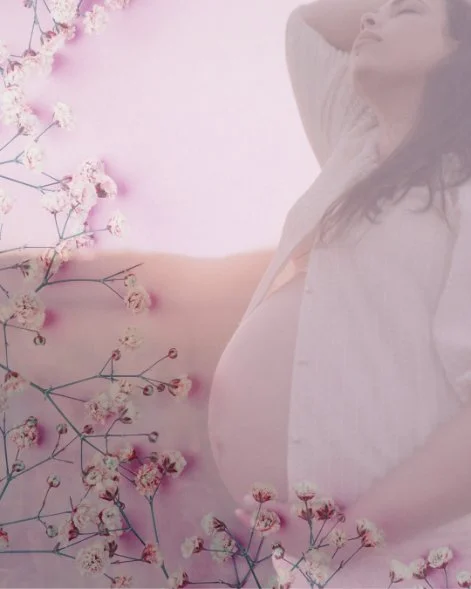 A pregnant woman relaxing with her eyes closed, wearing a white shirt, surrounded by pink flowers on a light background.