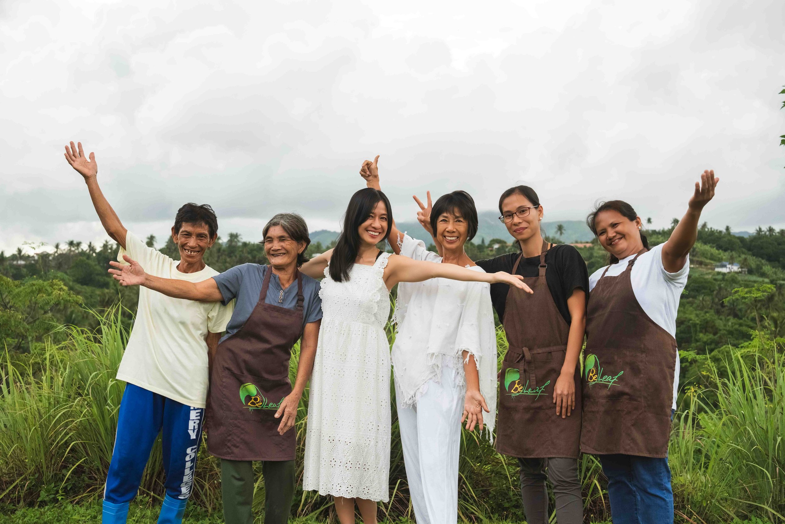 Group of six people standing outdoors in a field, smiling and making joyful gestures, with greenery and cloudy sky in the background.