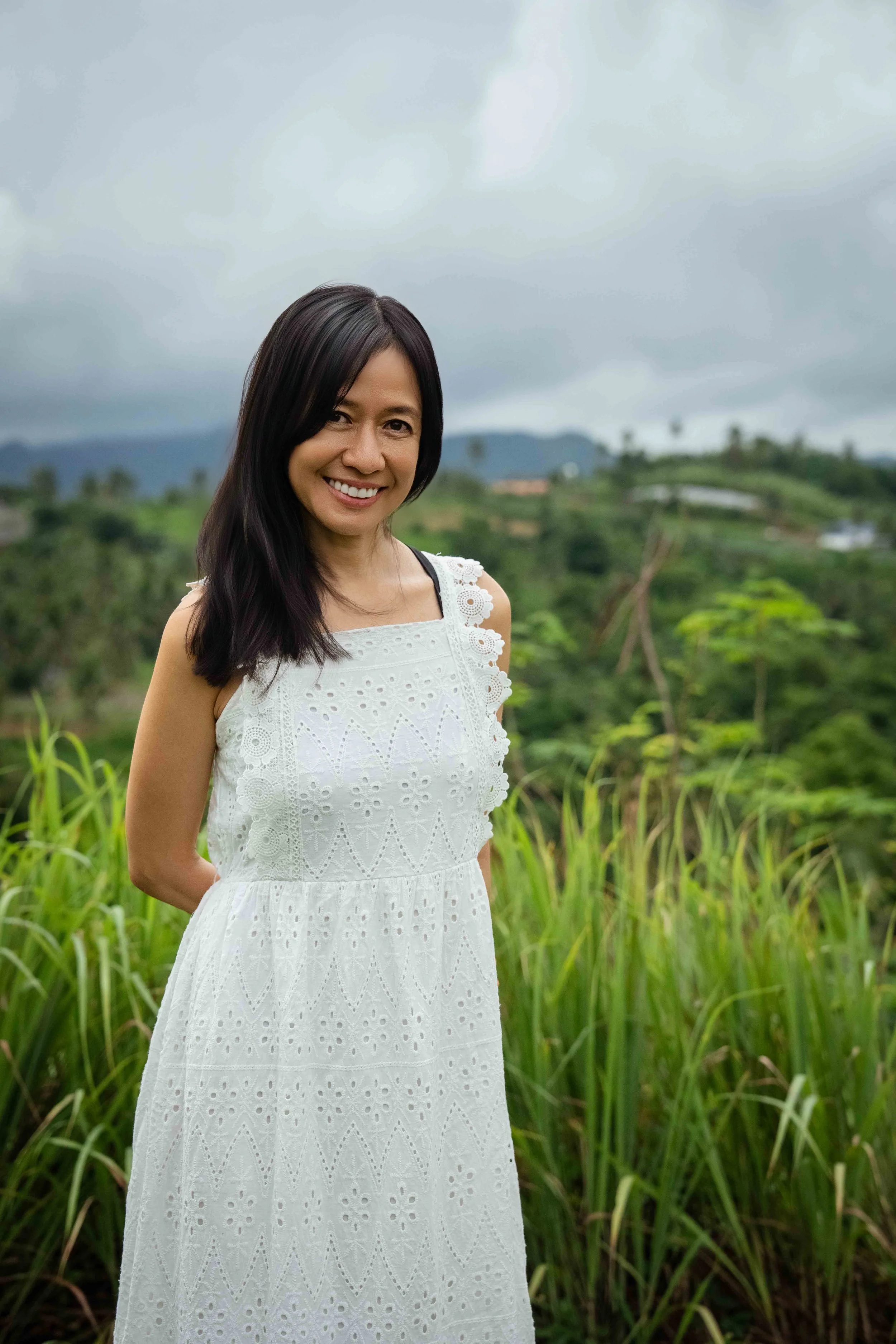 A woman in a white dress standing outdoors against a lush, green hilly landscape with cloudy sky.