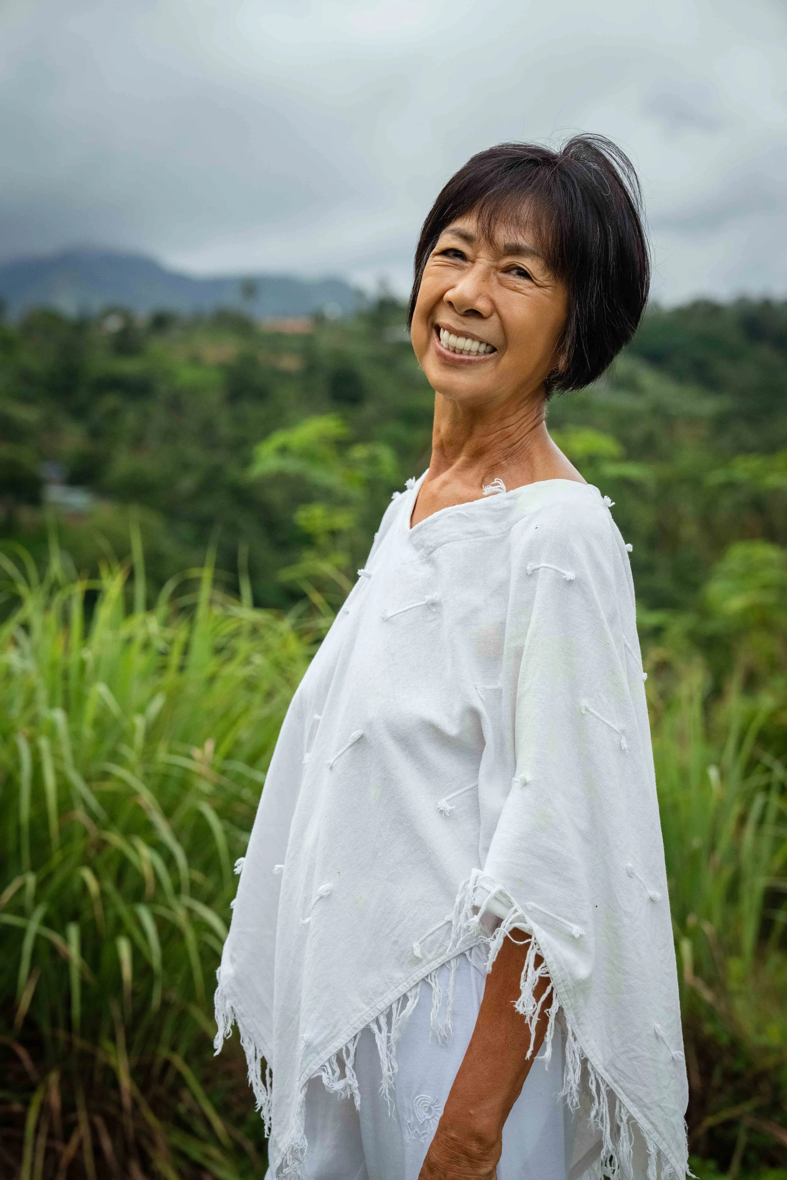 Smiling older woman with short black hair wearing a white, distressed, fringed top standing outdoors in a green, hilly landscape.