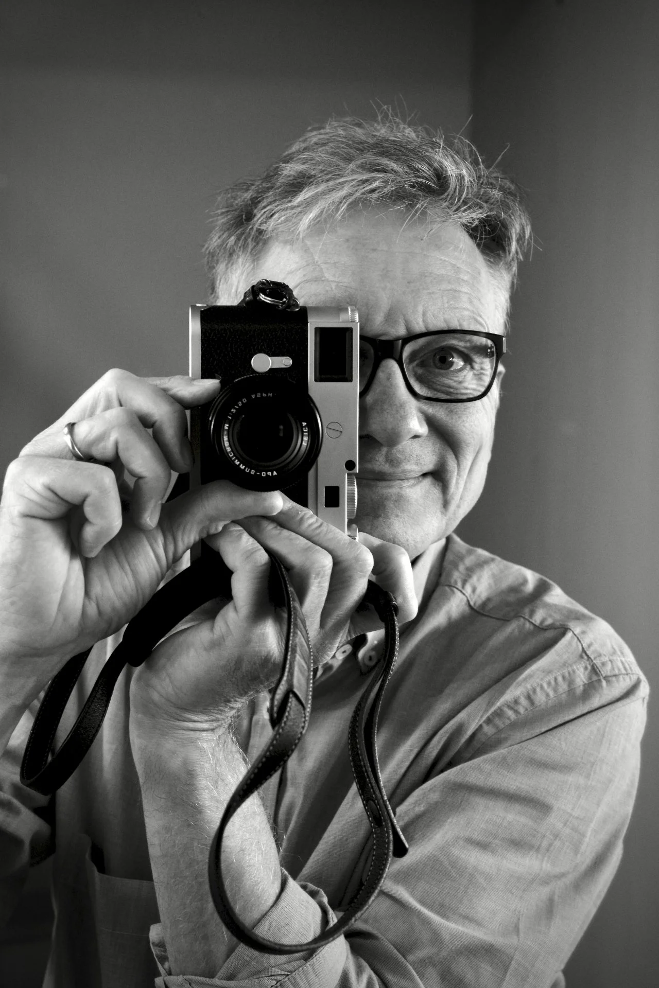 Ein Mann mit Brille und grauem Haar hält eine Kamera vor sein Gesicht und fotografiert sich selbst, während er in einen Spiegel schaut.