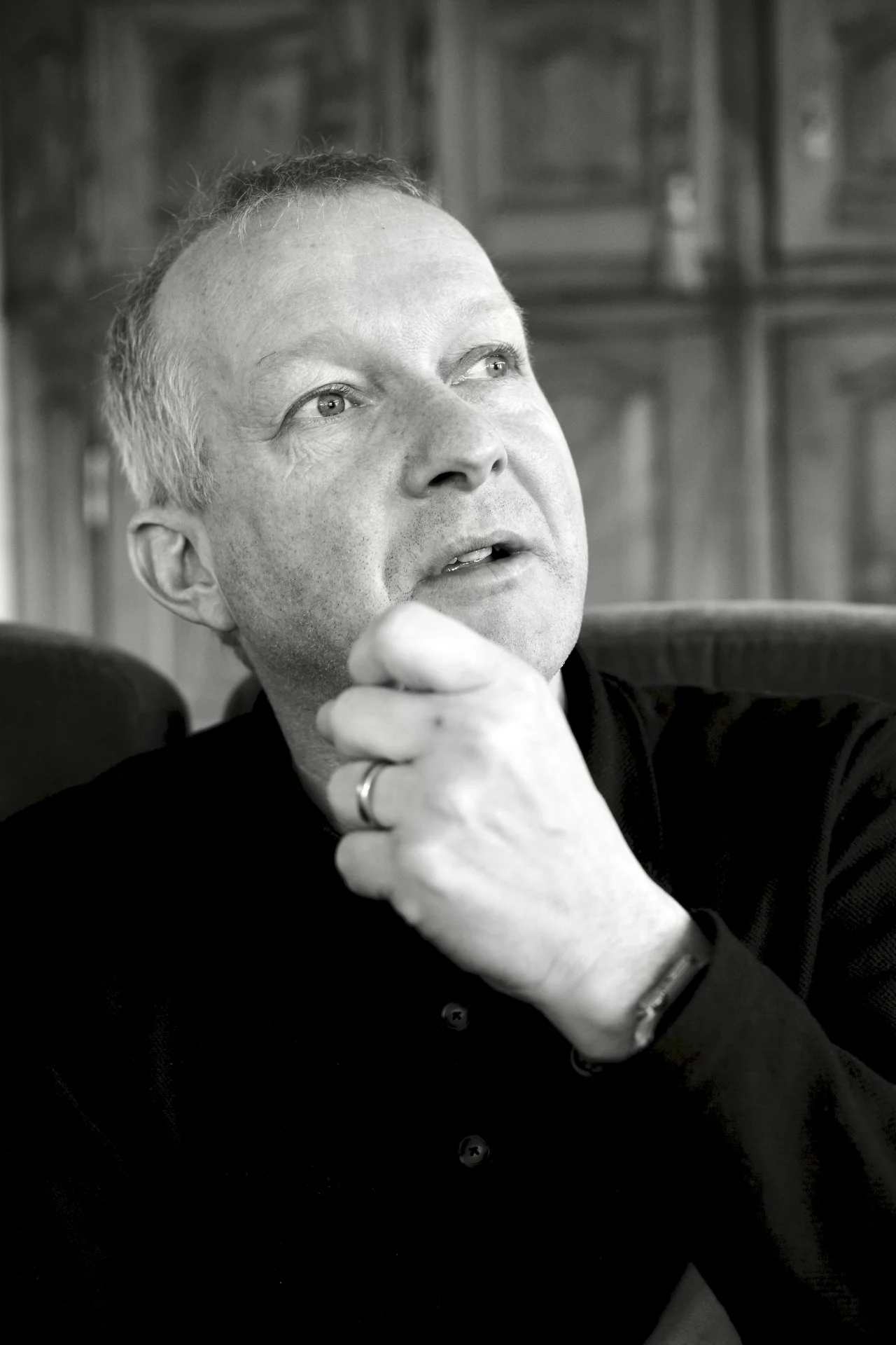 Schwarz-weiß-Foto eines Mannes, der mit nachdenklicher Miene seine Hand an sein Kinn legt, in einem Raum mit Holzmöbeln im Hintergrund.