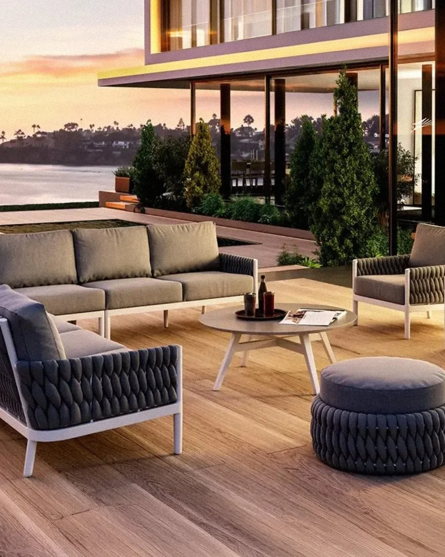 Outdoor patio with modern furniture, including a sofa, armchair, ottoman, and a small round table, overlooking a body of water with trees and houses in the background during sunset.