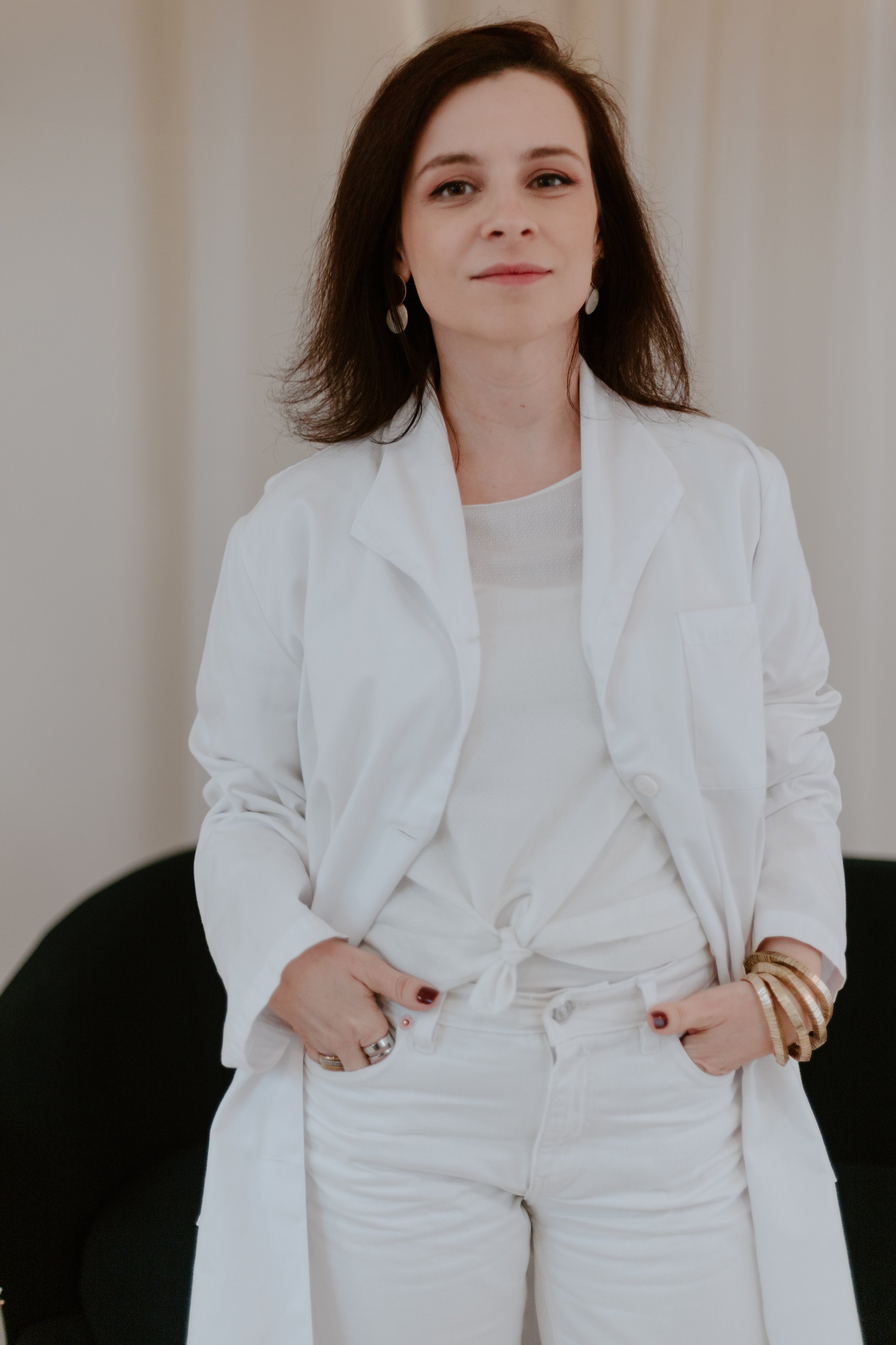 Mulher jovem com cabelo castanho, usando uma blusa branca e um casaco branco, de mãos nos bolsos, olhando para a câmera.