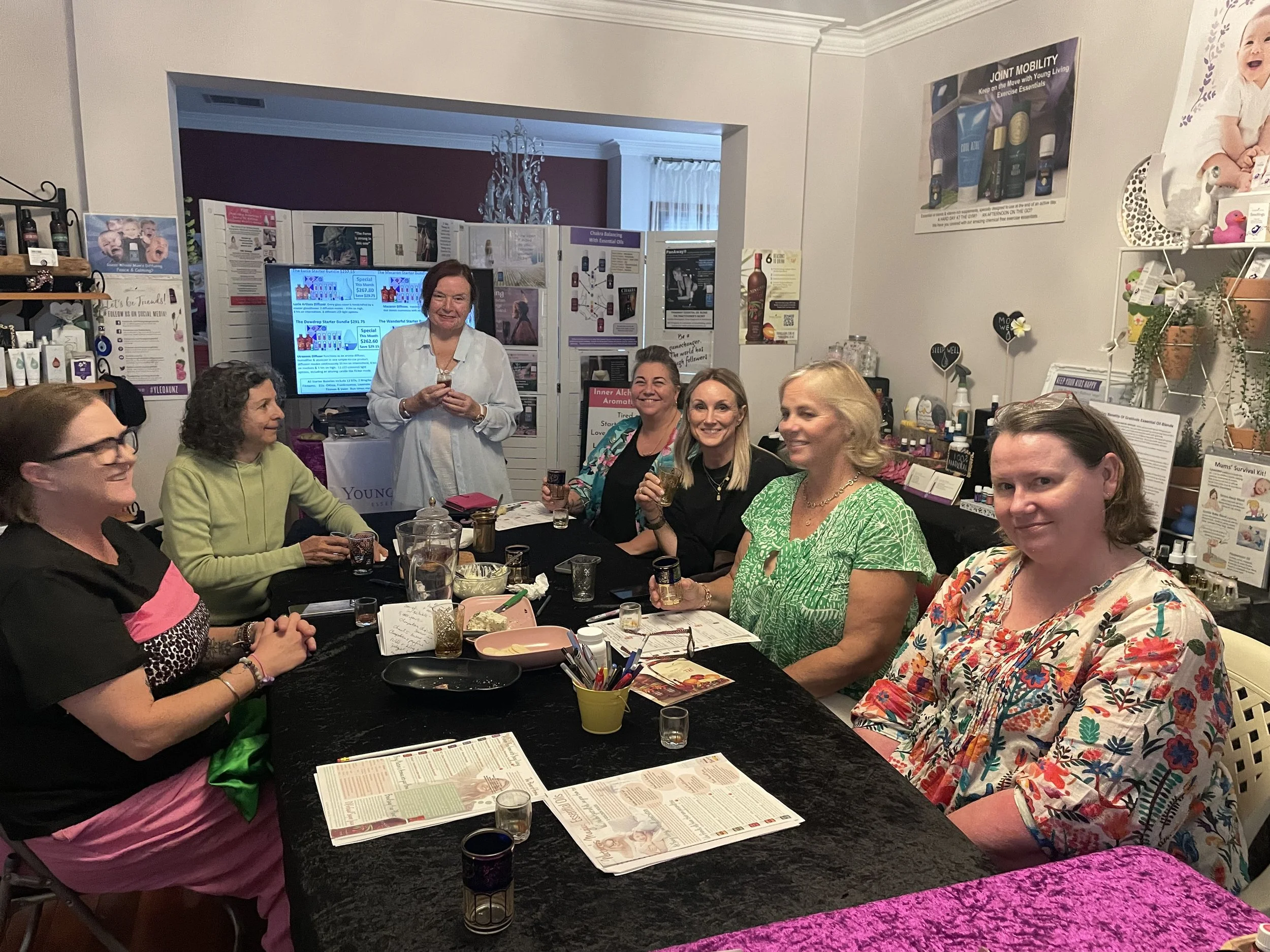 Group of women sitting around a table in a room with a screen and posters, celebrating with drinks and smiling at the camera.