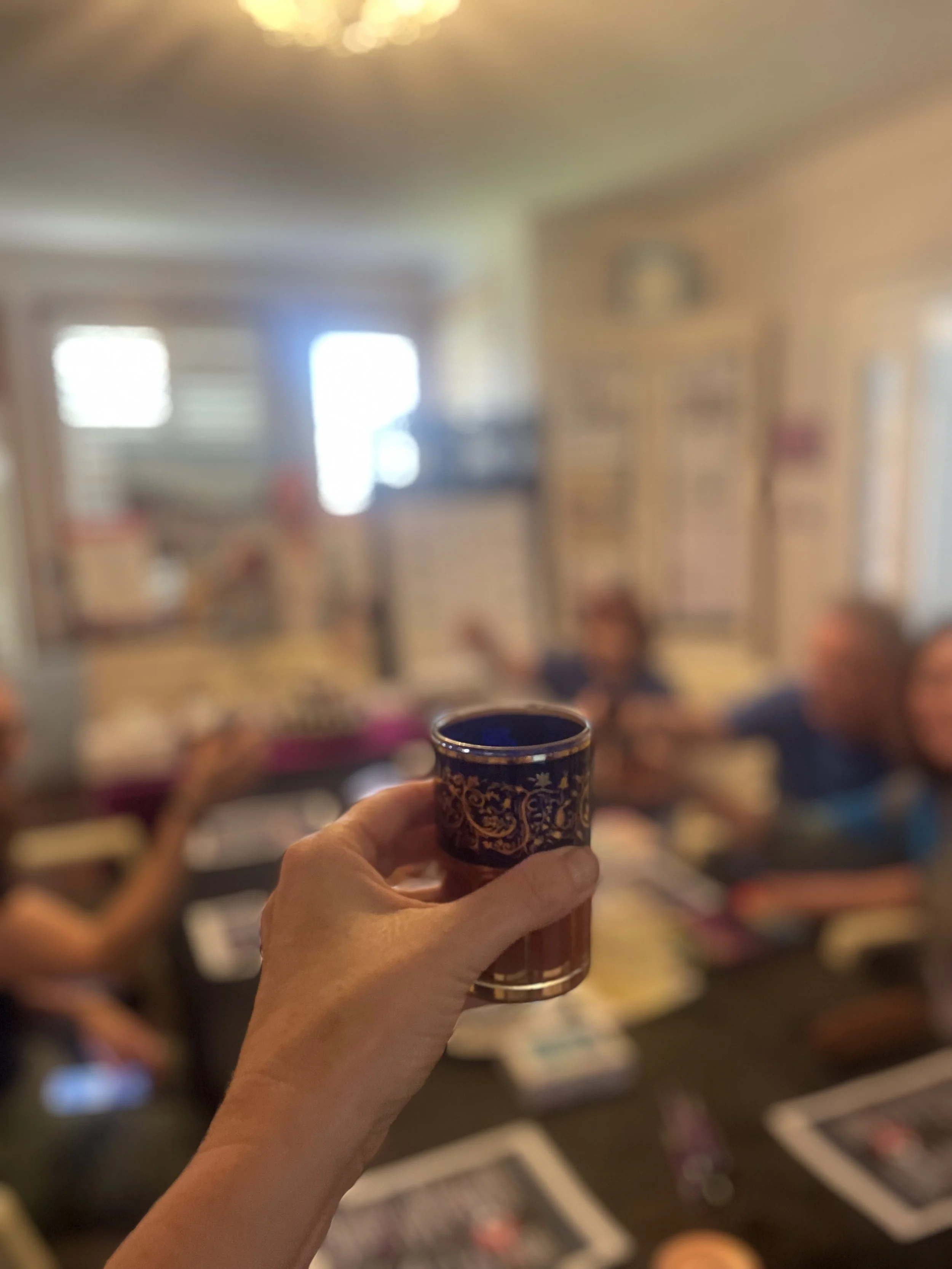 A hand holds a small ornate glass cup with a pattern, in focus. In the background, there are people around a table with tablets and books, in a cozy, well-lit room with windows.