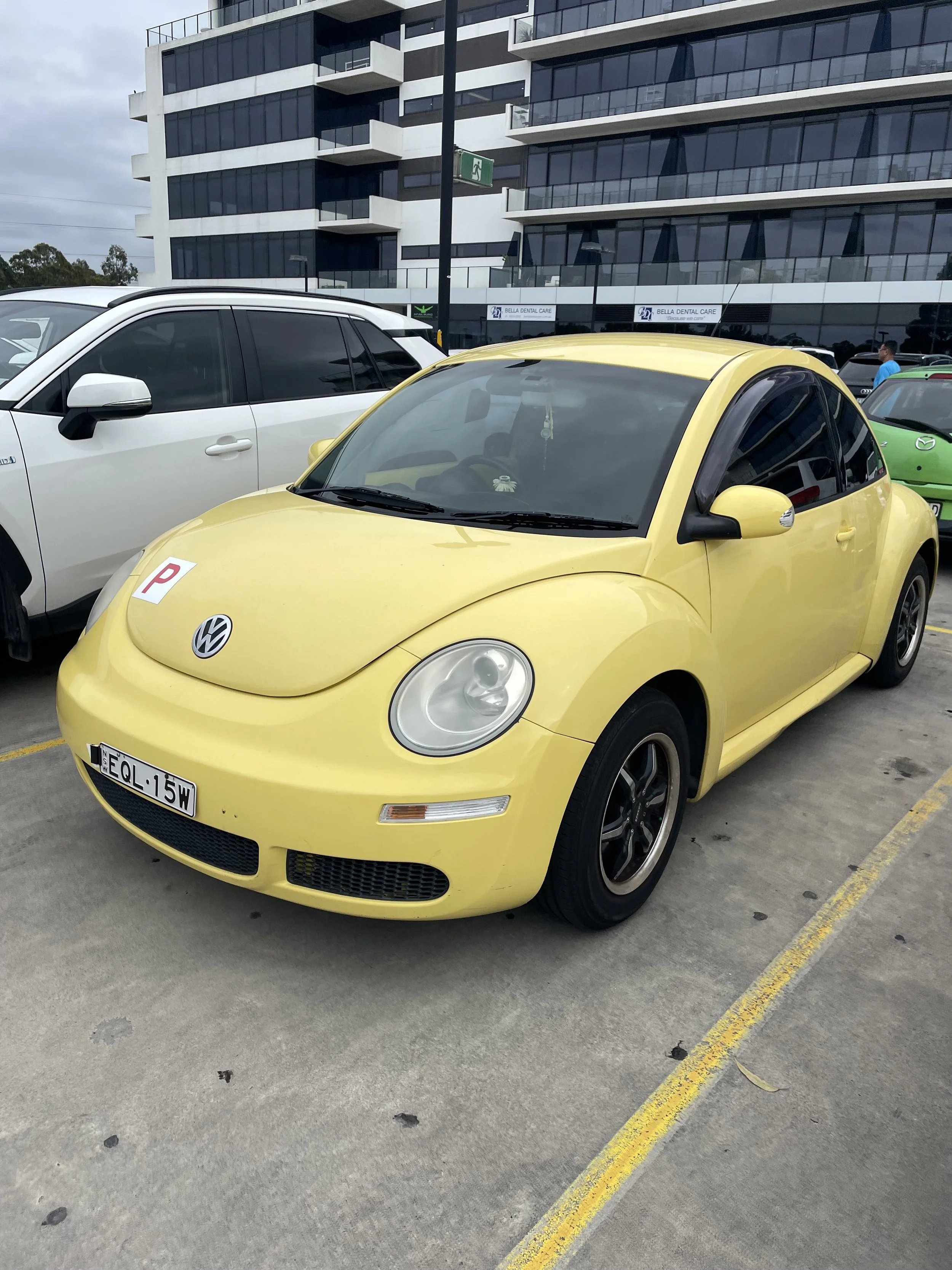 Yellow Volkswagen Beetle parked in a parking lot next to white and green cars, with a modern building in the background.