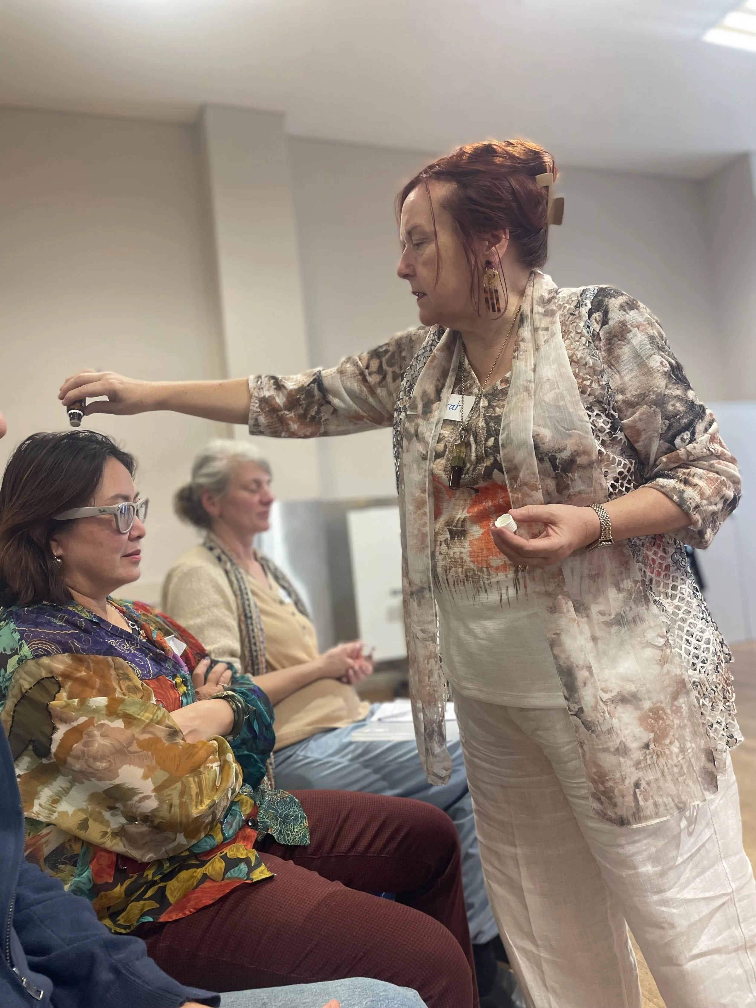 A woman with red hair is holding an essential oil bottle above a seated woman with glasses, who is looking up at her. Two other women are seated nearby, listening attentively.