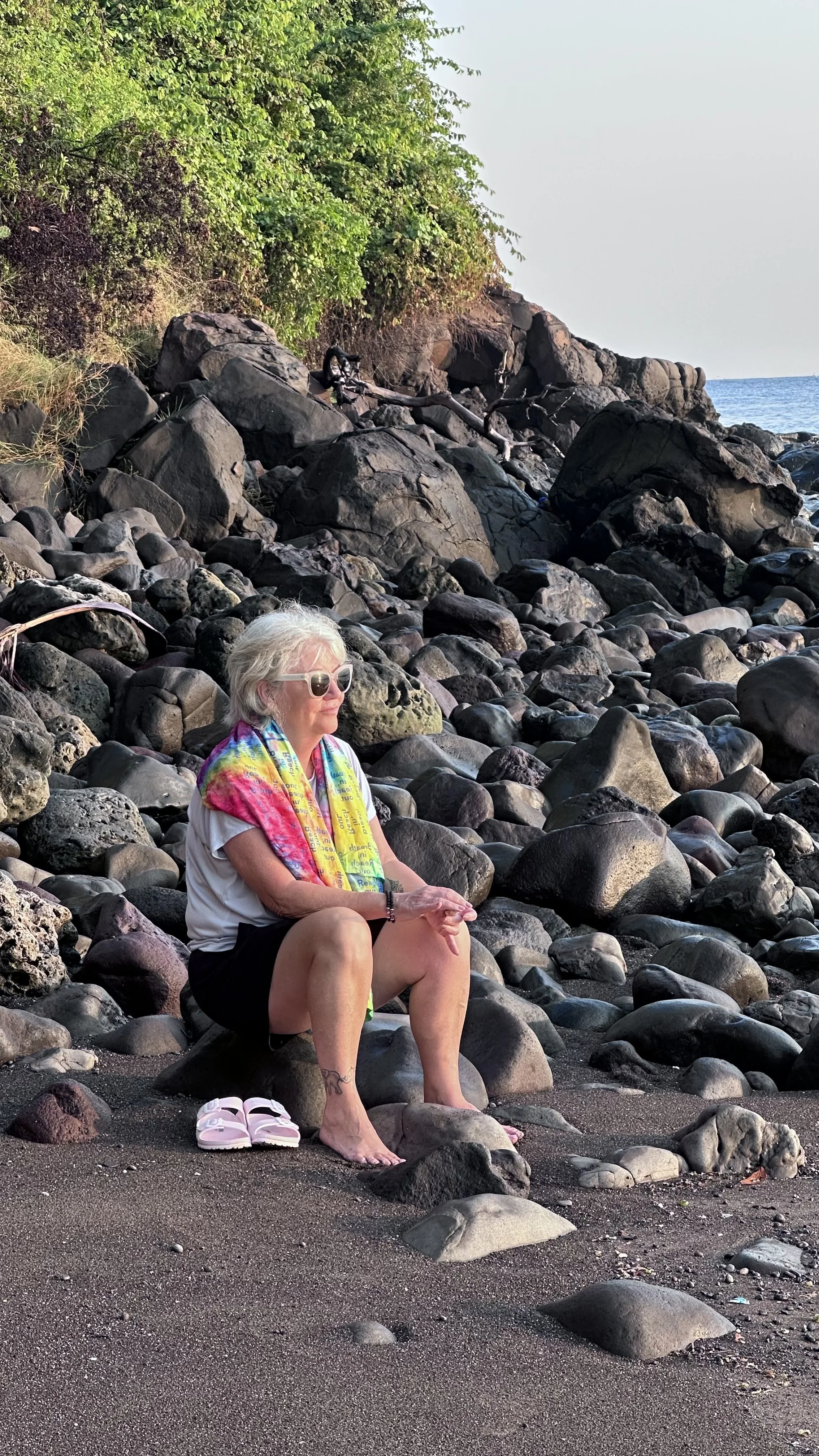 An elderly woman with white hair, wearing sunglasses, a colorful scarf, a white t-shirt, and black shorts, sitting on a rock on a rocky beach, with her shoes placed beside her. She appears to be meditating or relaxing while facing the ocean.