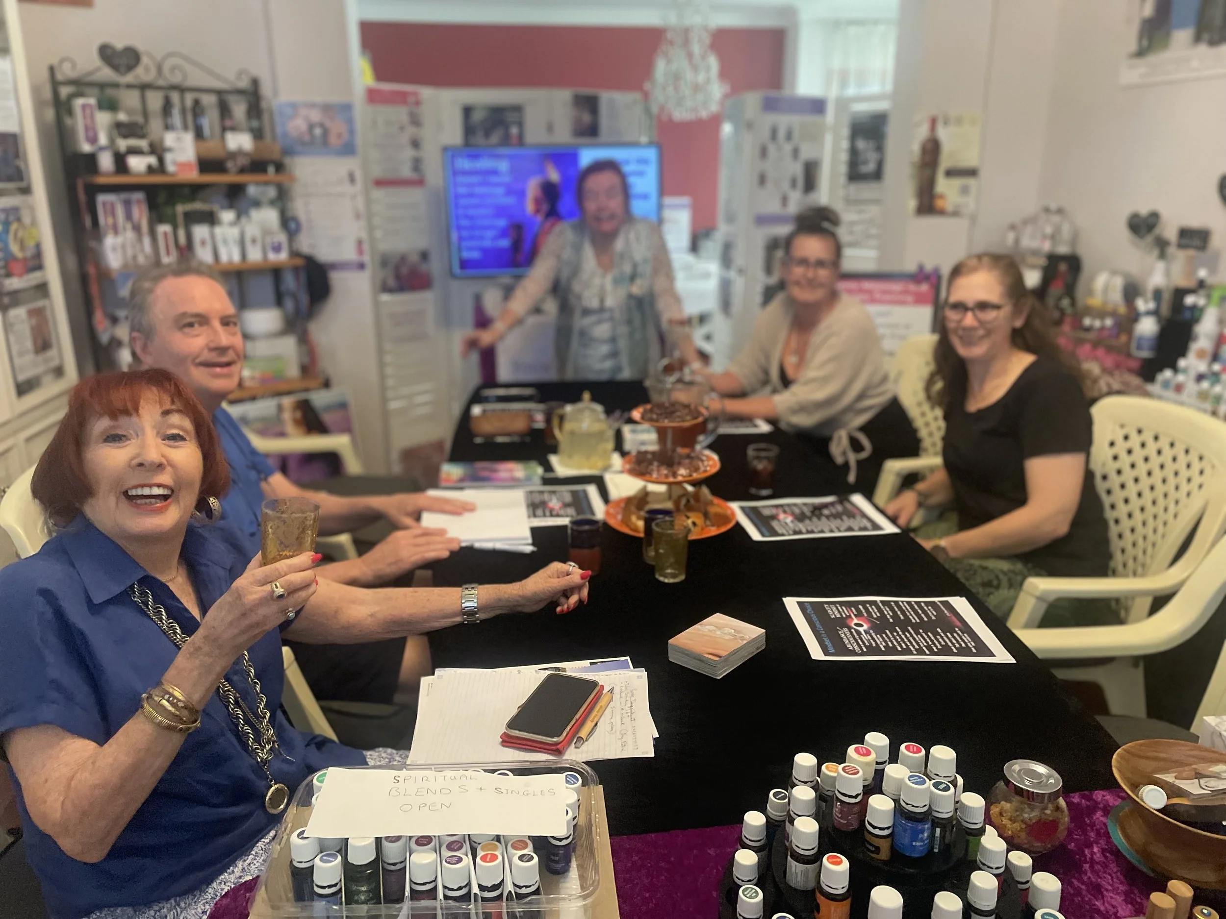 A group of five women sitting around a table, smiling and holding drinks, with a display of essential oils and wellness products on the table in a cozy room decorated with shelves, posters, and a television in the background.