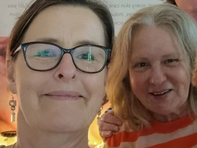 Two women smiling and posing for a photo, one with glasses and the other with shoulder-length blonde hair, in an indoor setting with warm lighting.