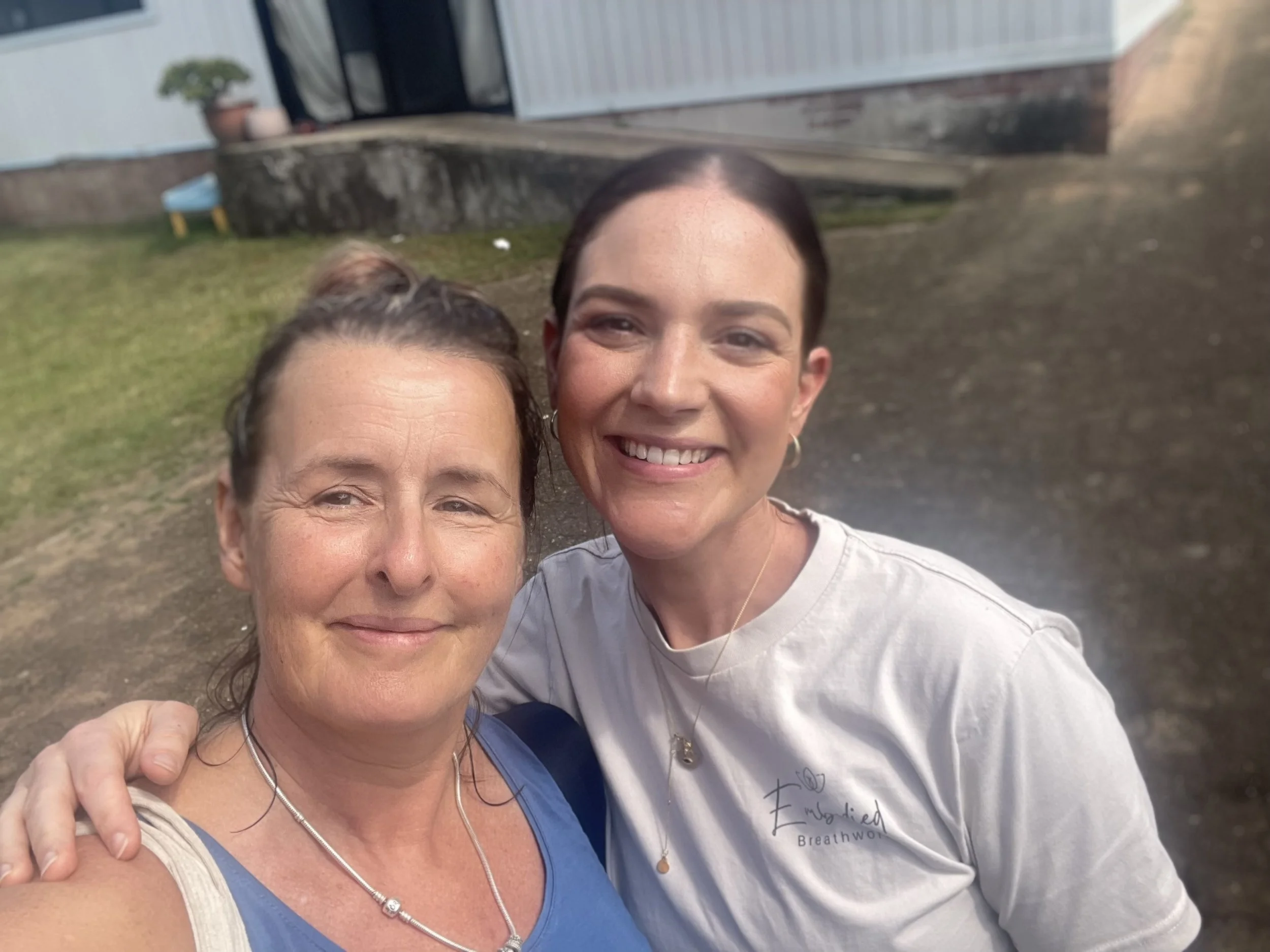 Two women smiling outdoors, one with dark hair in a ponytail, wearing a blue top and necklace, and the other with dark hair in a bun, wearing a light gray shirt with text, standing close together in front of a grassy yard with a brick and white building in the background.
