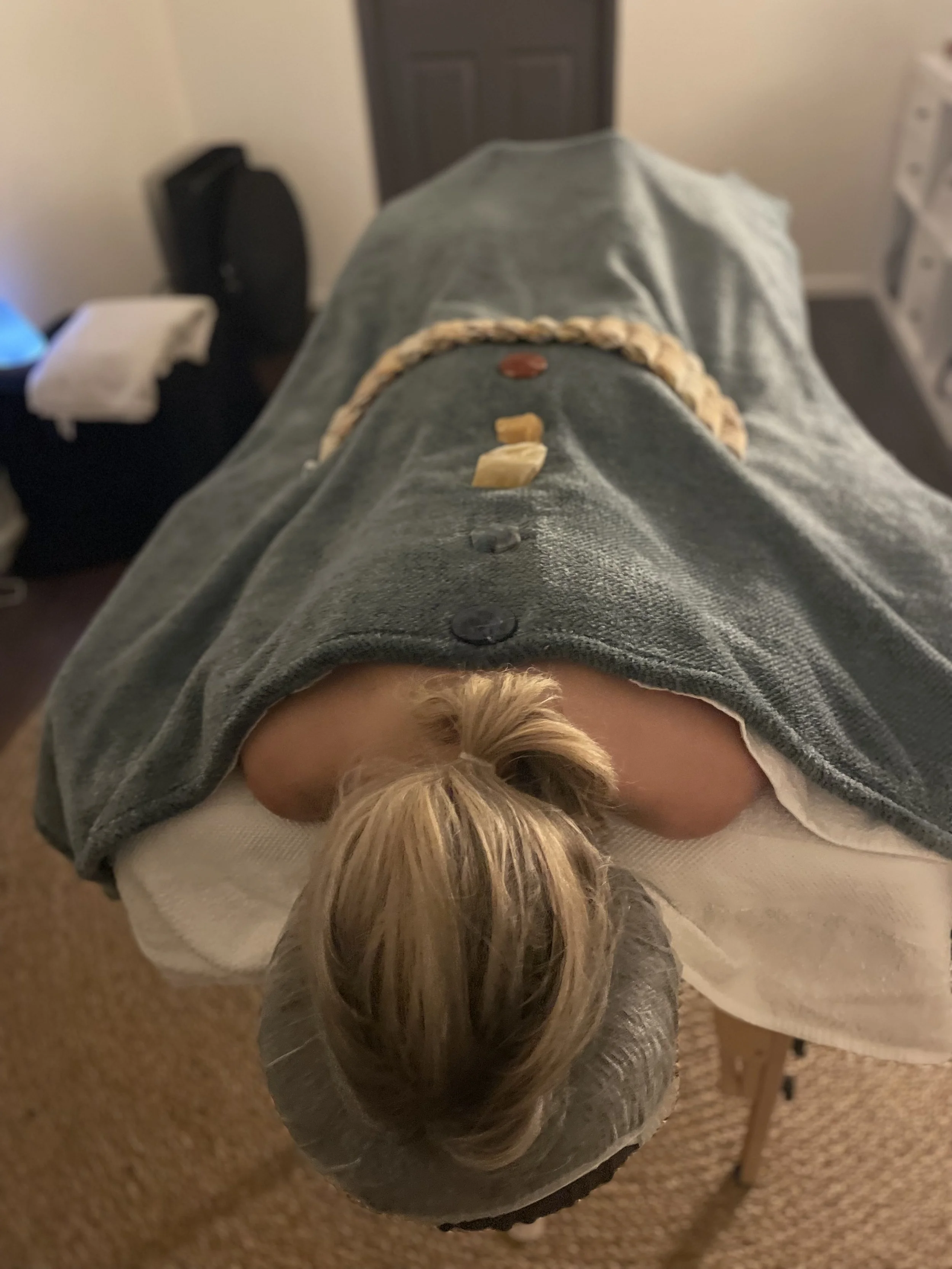 A person receiving a massage lying face down on a massage table, covered with a gray towel, with stones and herbs placed on their back.