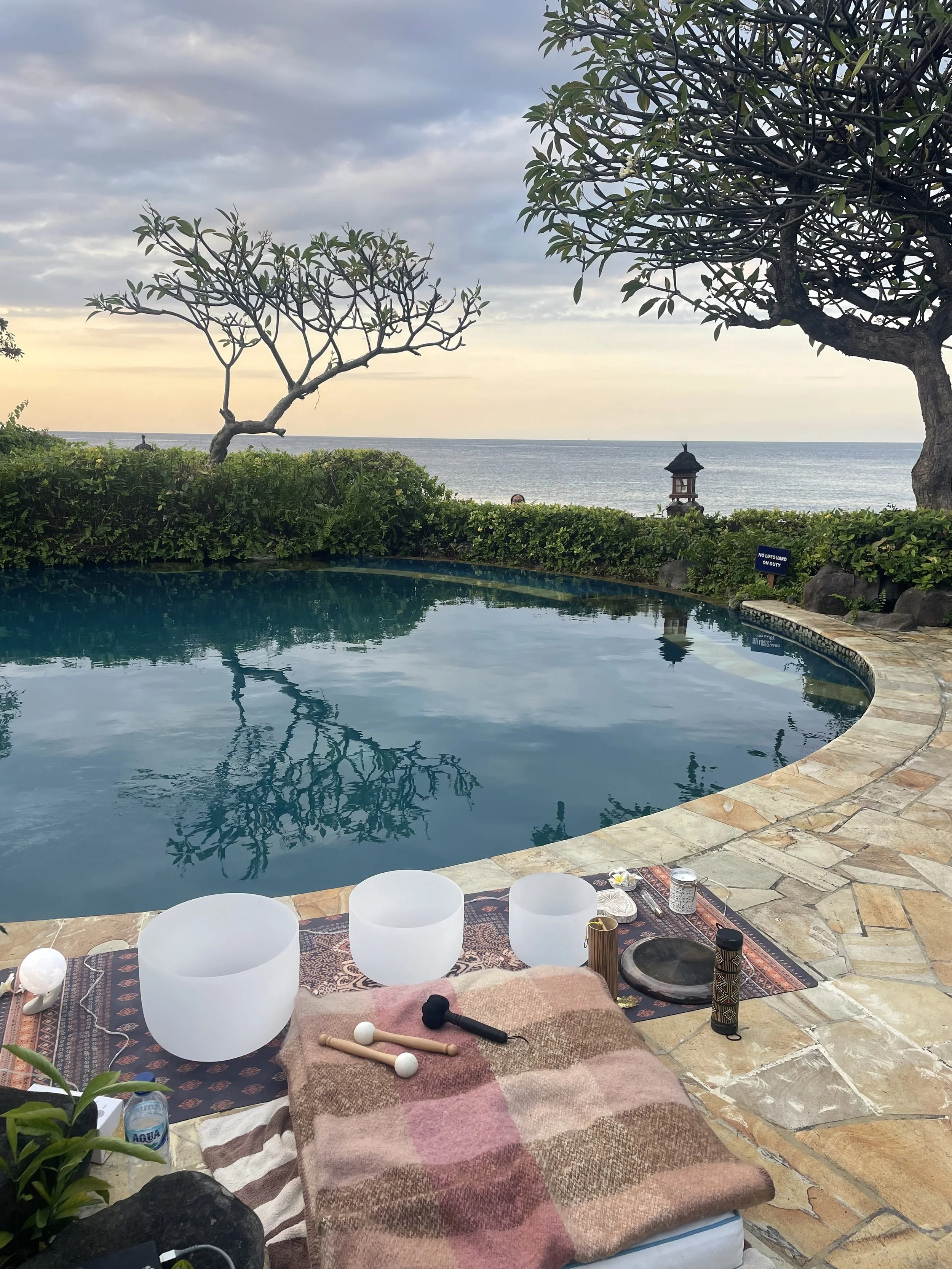 A tranquil outdoor pool with a seaside view, surrounded by greenery and trees, featuring a mat with crystal singing bowls, mallets, and various accessories.