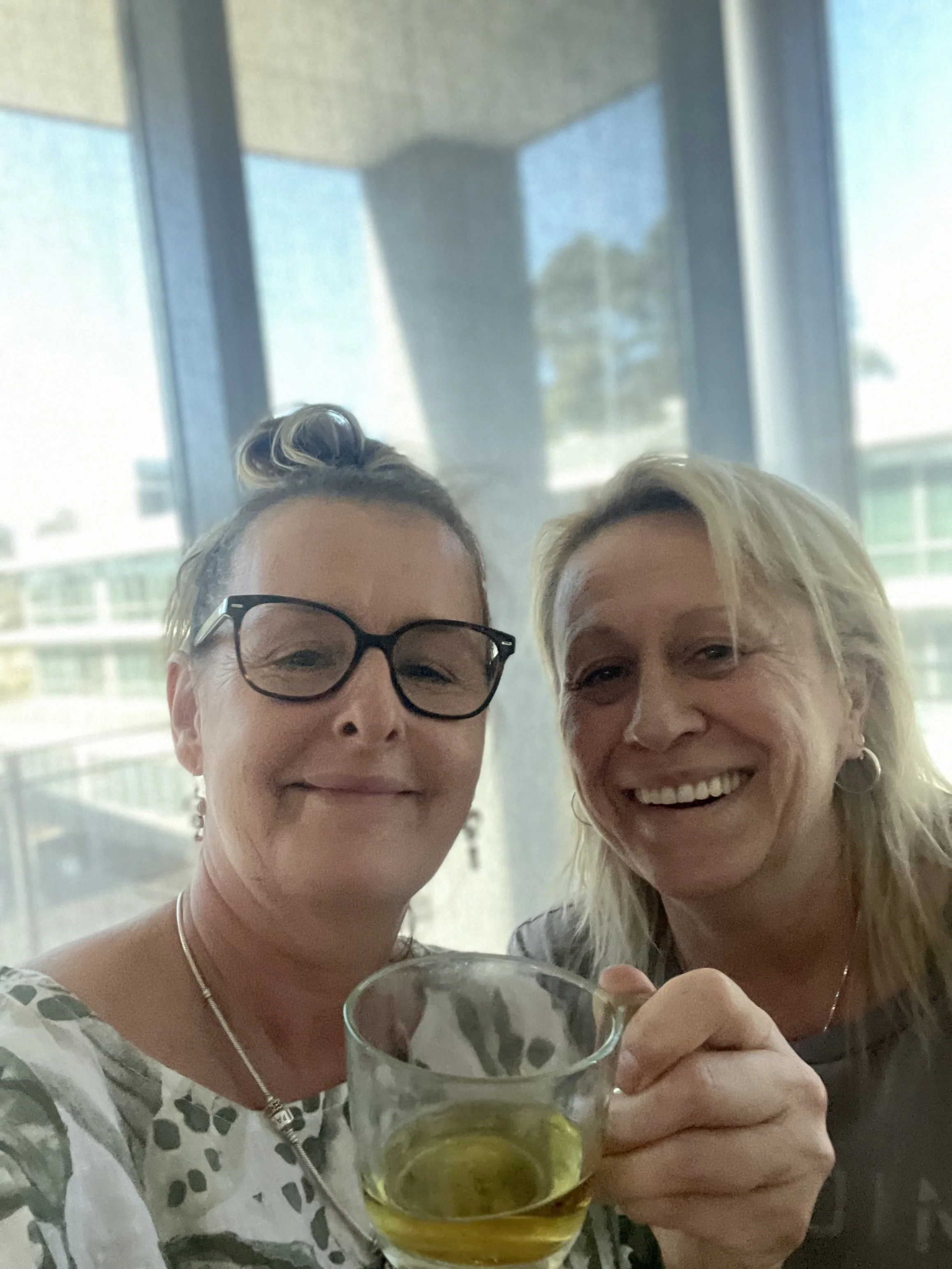 Two women smiling, one holding a glass of drink, in front of large windows with a cityscape background.