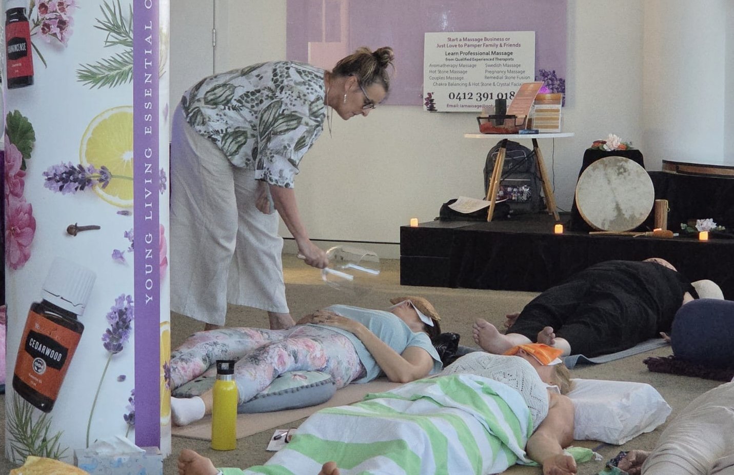 A woman performing a guided meditation or relaxation session for a group of people lying on the floor with their eyes closed. The space appears to be a wellness or spa setting with relaxing decor and essential oils.