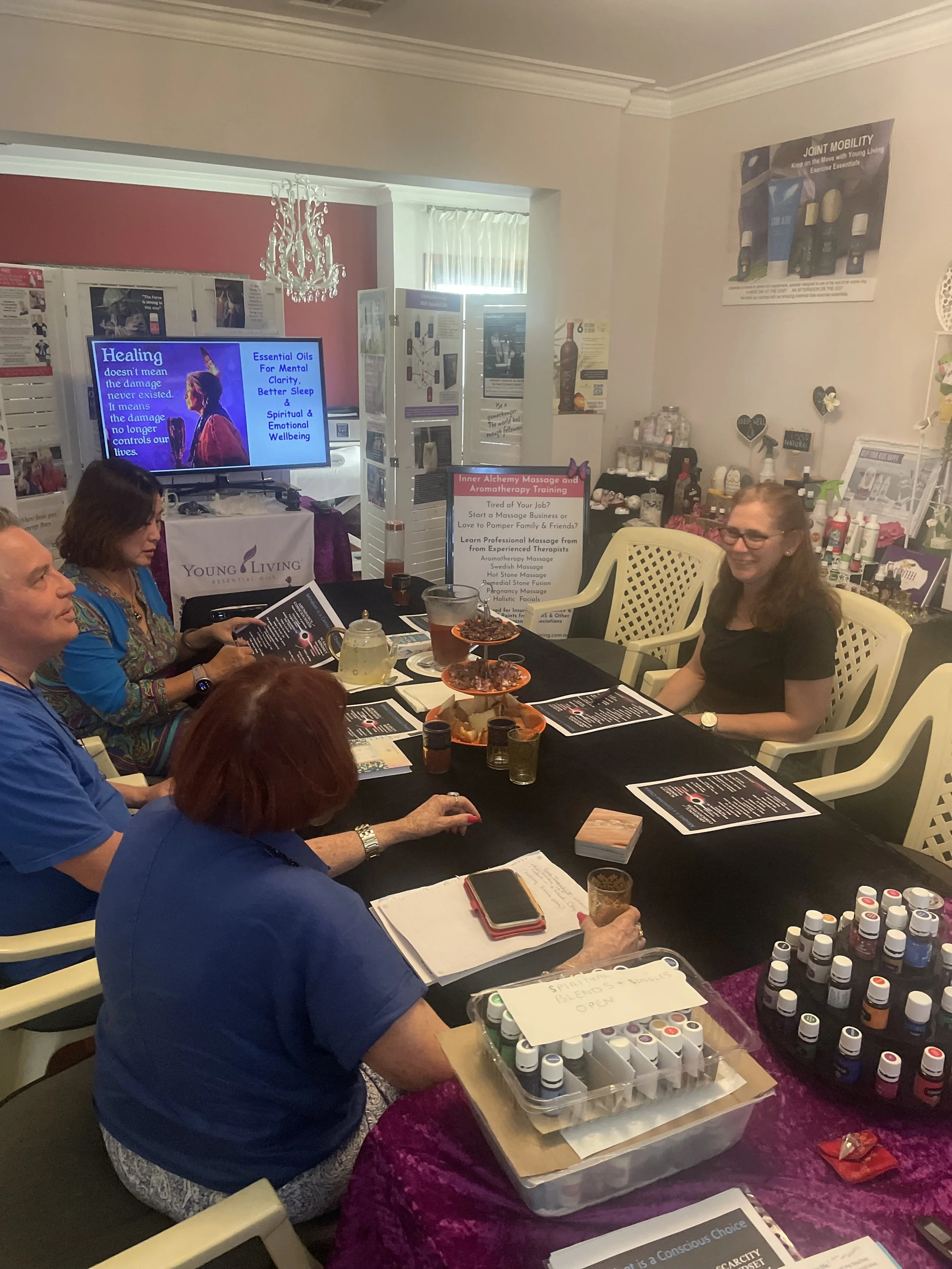 A group of people sitting around a table during a workshop or seminar, with various informational materials, essential oils, and a large screen in the background displaying a presentation about healing and essential oils.
