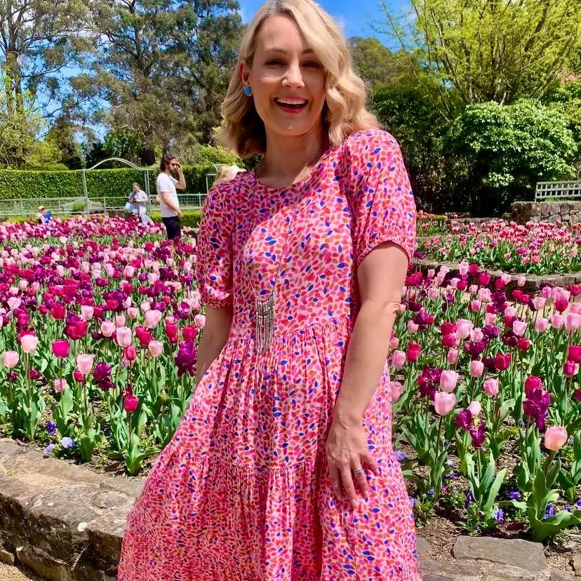 A woman with blonde hair smiling and wearing a pink patterned dress in a colorful tulip garden.