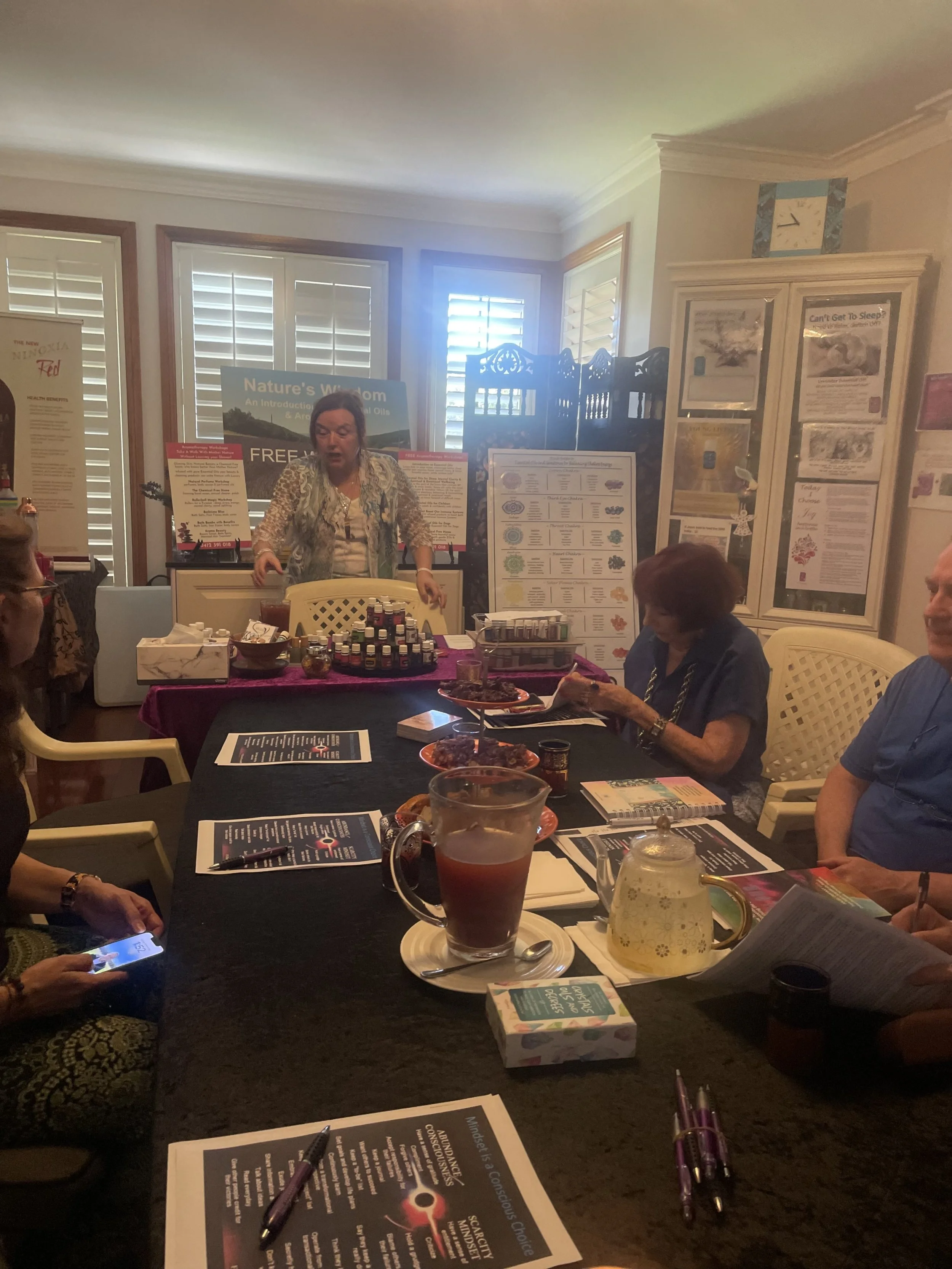 People sitting around a table with informational pamphlets, a pitcher of tea, and some snacks, while a woman explains various products related to essential oils at a presentation or workshop inside a room with windows and informational posters on the