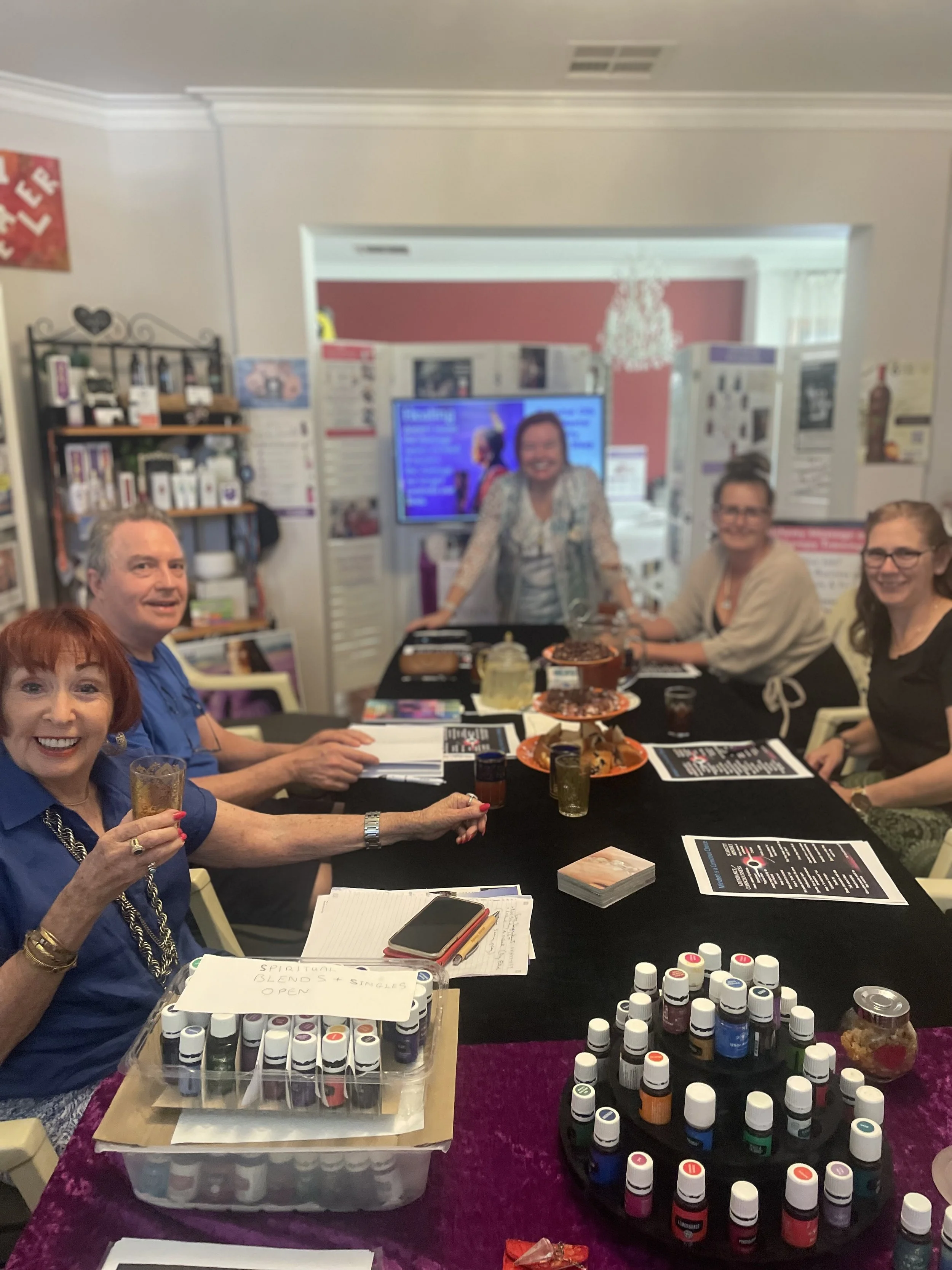 Group of five women sitting around a table at a social gathering, with various bottles of essential oils, notepads, and drinks on the table, in a room with decorations and a television in the background.