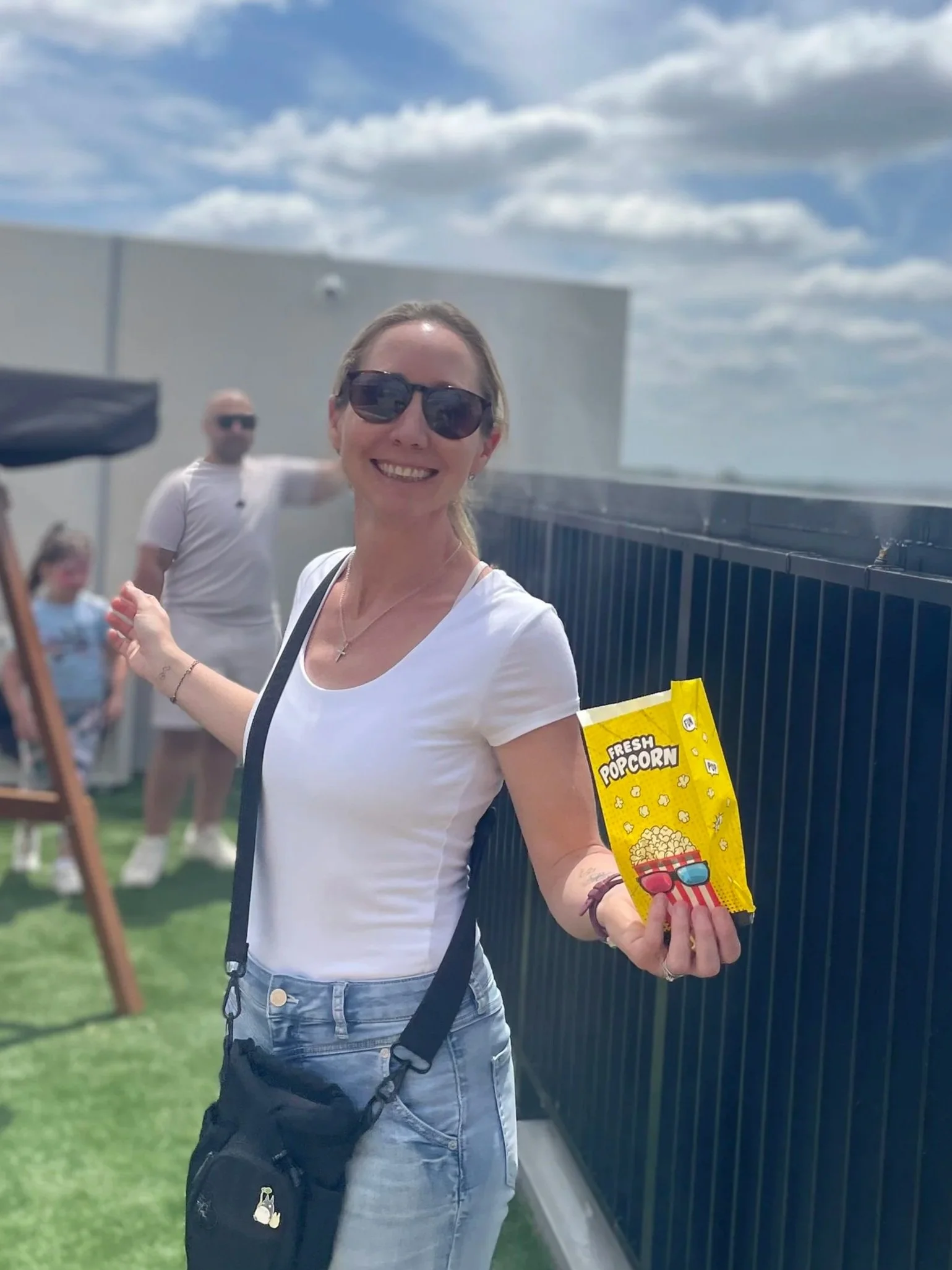 Smile woman wearing sunglasses holding a small bag of popcorn at an outdoor event during daytime.