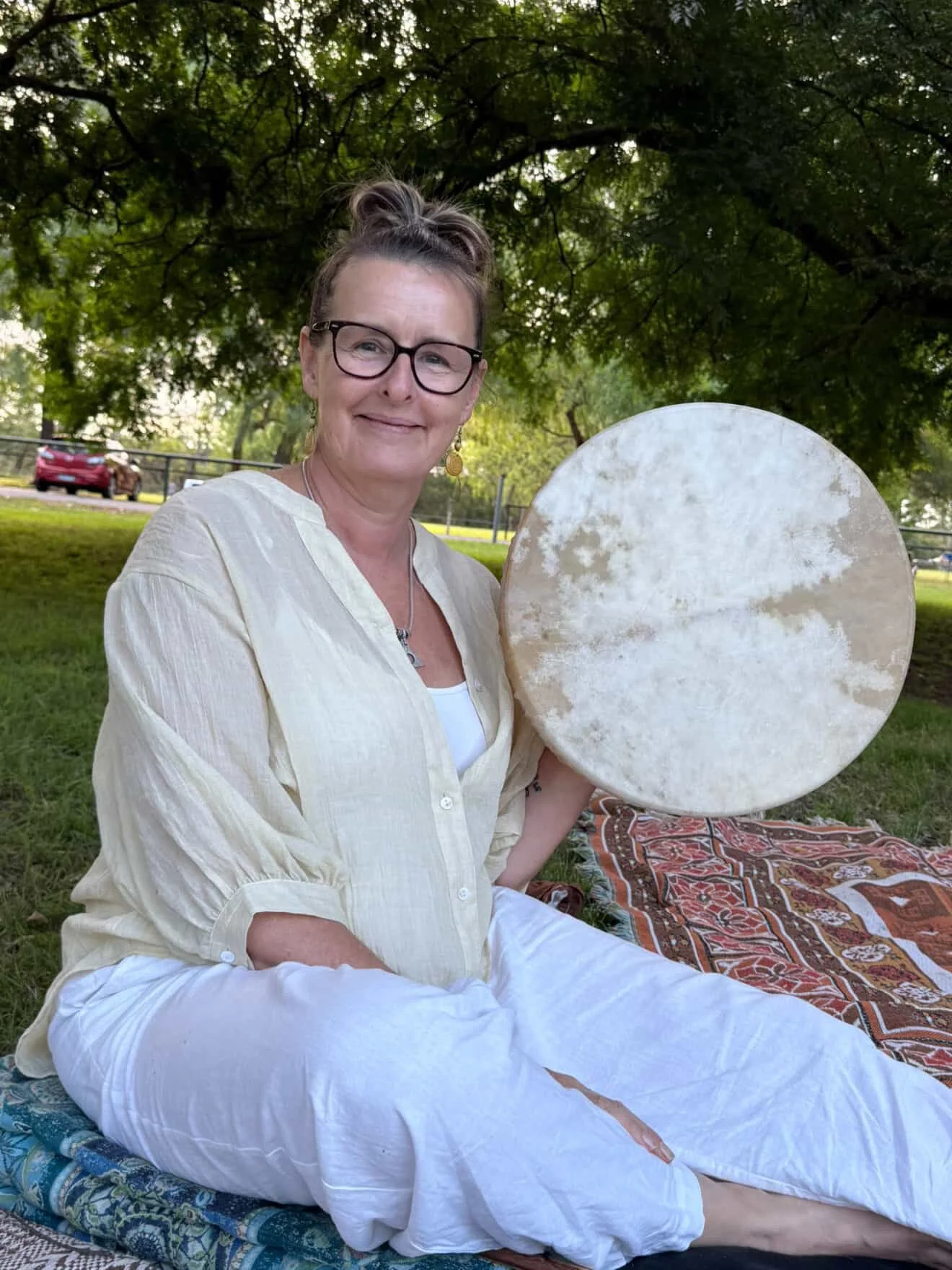 A woman with glasses sitting on a blanket outdoors holding a large, round white drum.