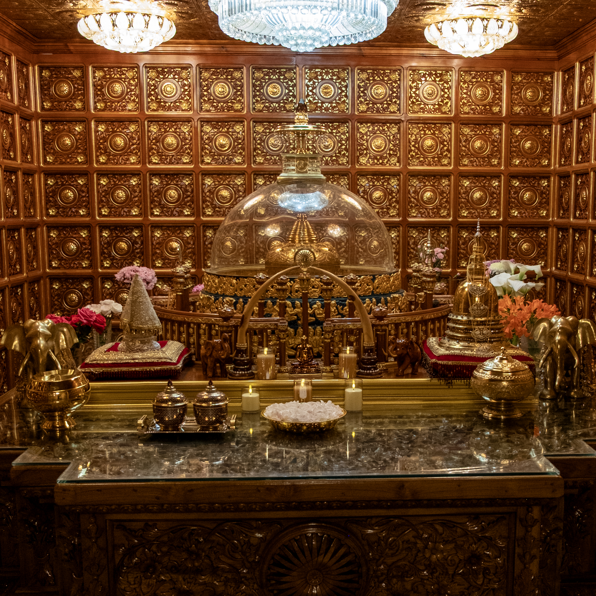 A richly decorated wooden altar with gold accents, adorned with candles, flowers, silver and gold vessels, and small statues. The background features a carved wooden wall, and chandeliers hang from the ceiling.