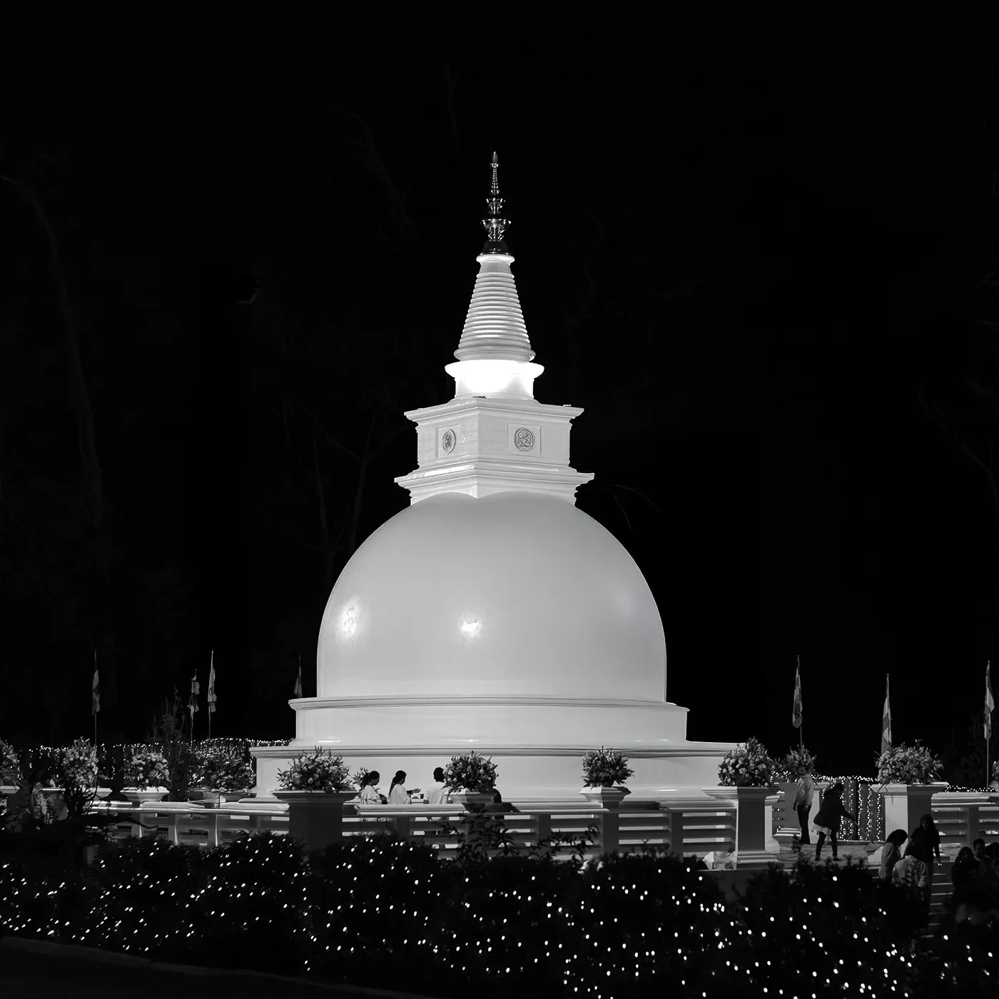 Nighttime view of a white stupa with a large dome, surrounded by flowers and small lights, against a dark sky.