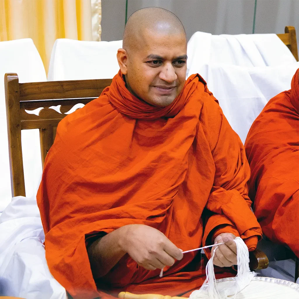 A Buddhist monk with a shaved head, wearing traditional orange robes, sitting indoors, holding white string or thread in his hand.
