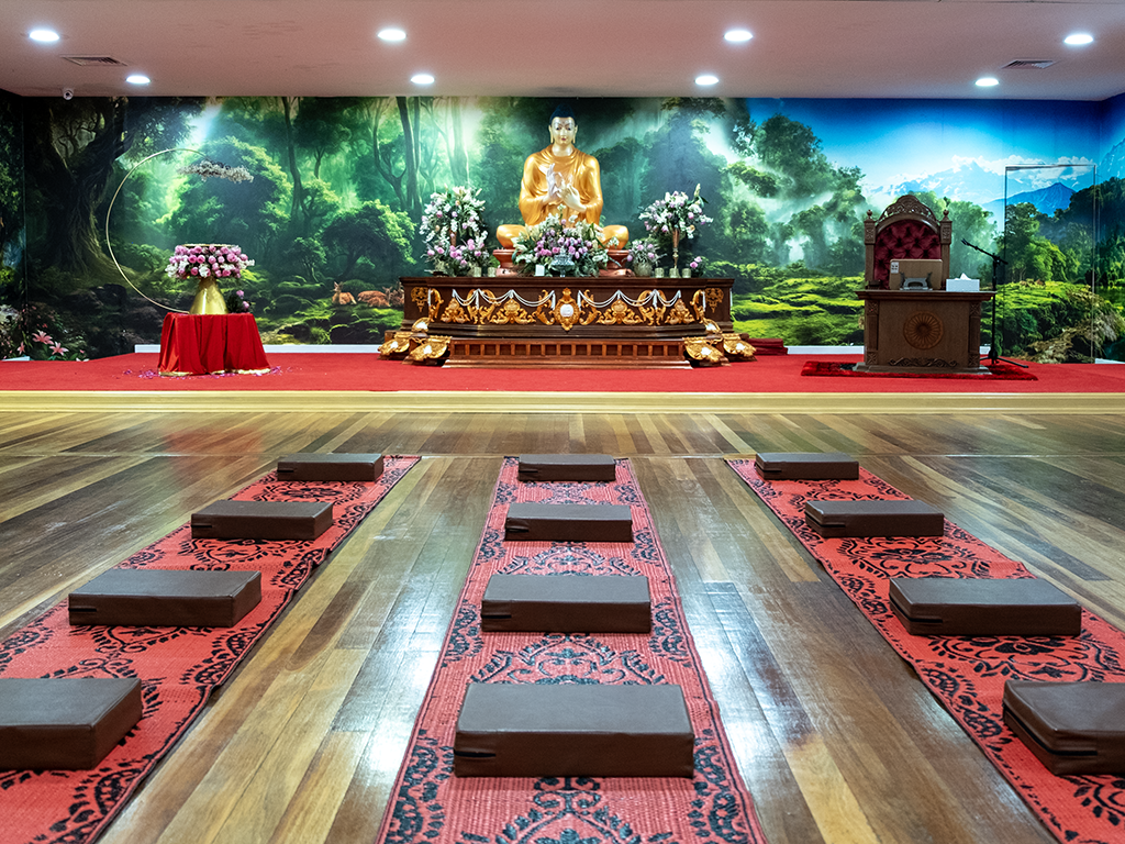 A meditation or prayer room set up with cushions and mats on a wooden floor, an altar with a statue of Buddha, and a scenic forest mural on the back wall.