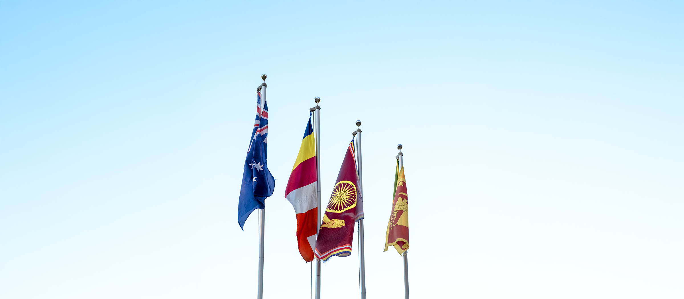 Four flags on flagpoles against a clear blue sky, including the Australian flag, the Ukrainian flag, the Sri Lankan flag, and the Sri Lankan flag.